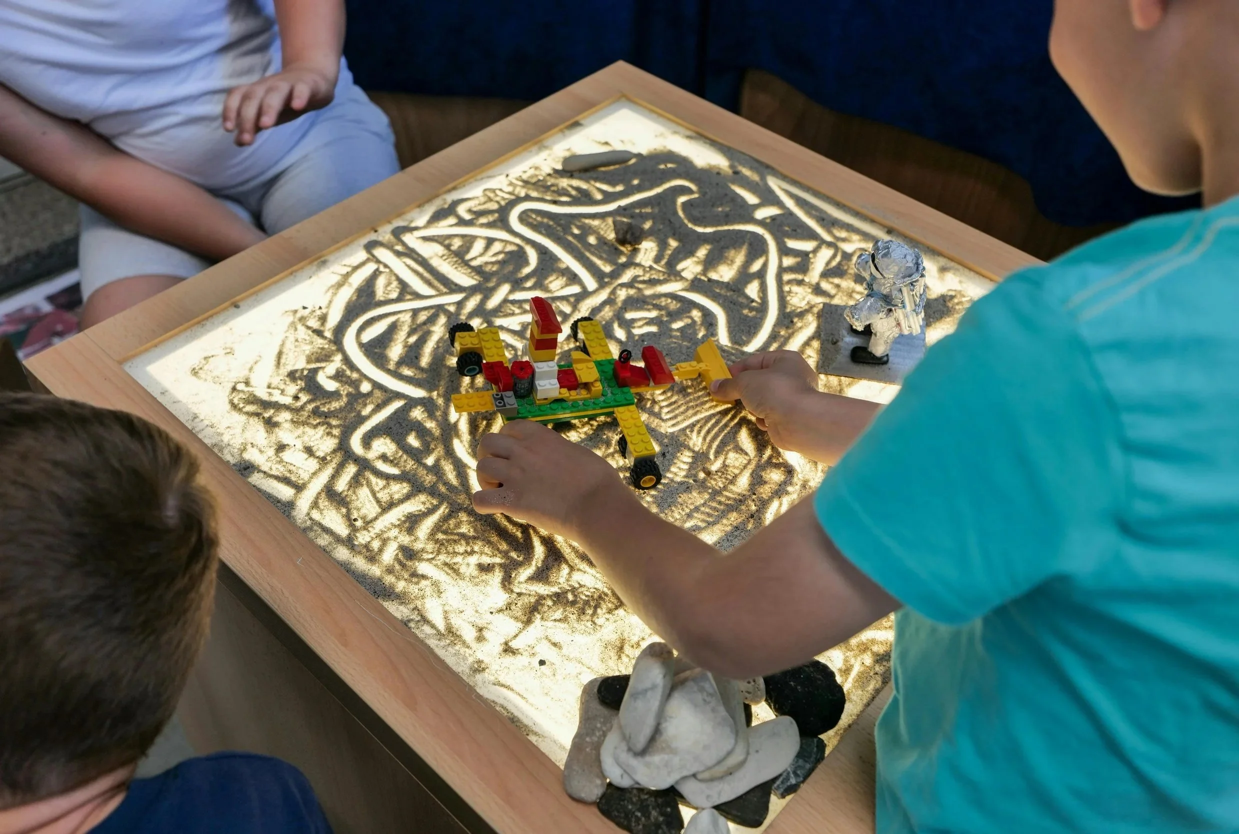Children playing with LEGO and toy astronaut figurine on a backlit sand tray with rocks.