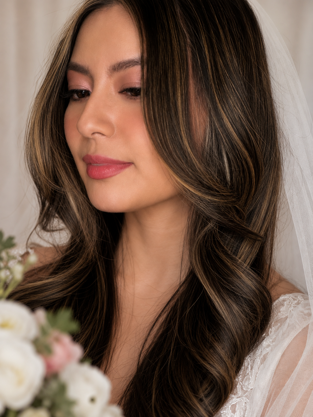 Close-up of a woman with long, wavy brown hair with highlights, wearing a wedding dress, and a white veil, holding a bouquet of white and pink flowers, in a soft-focus indoor setting.