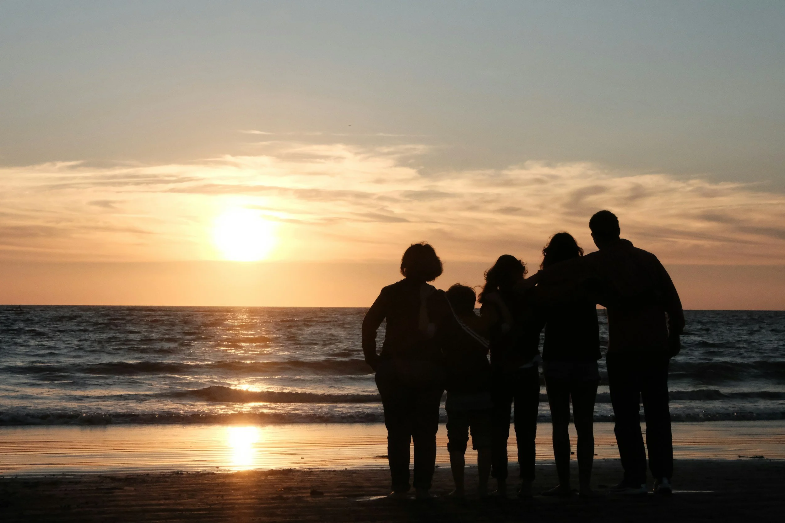 Group of five people standing on a beach at sunset, facing the ocean and watching the sun descend below the horizon.