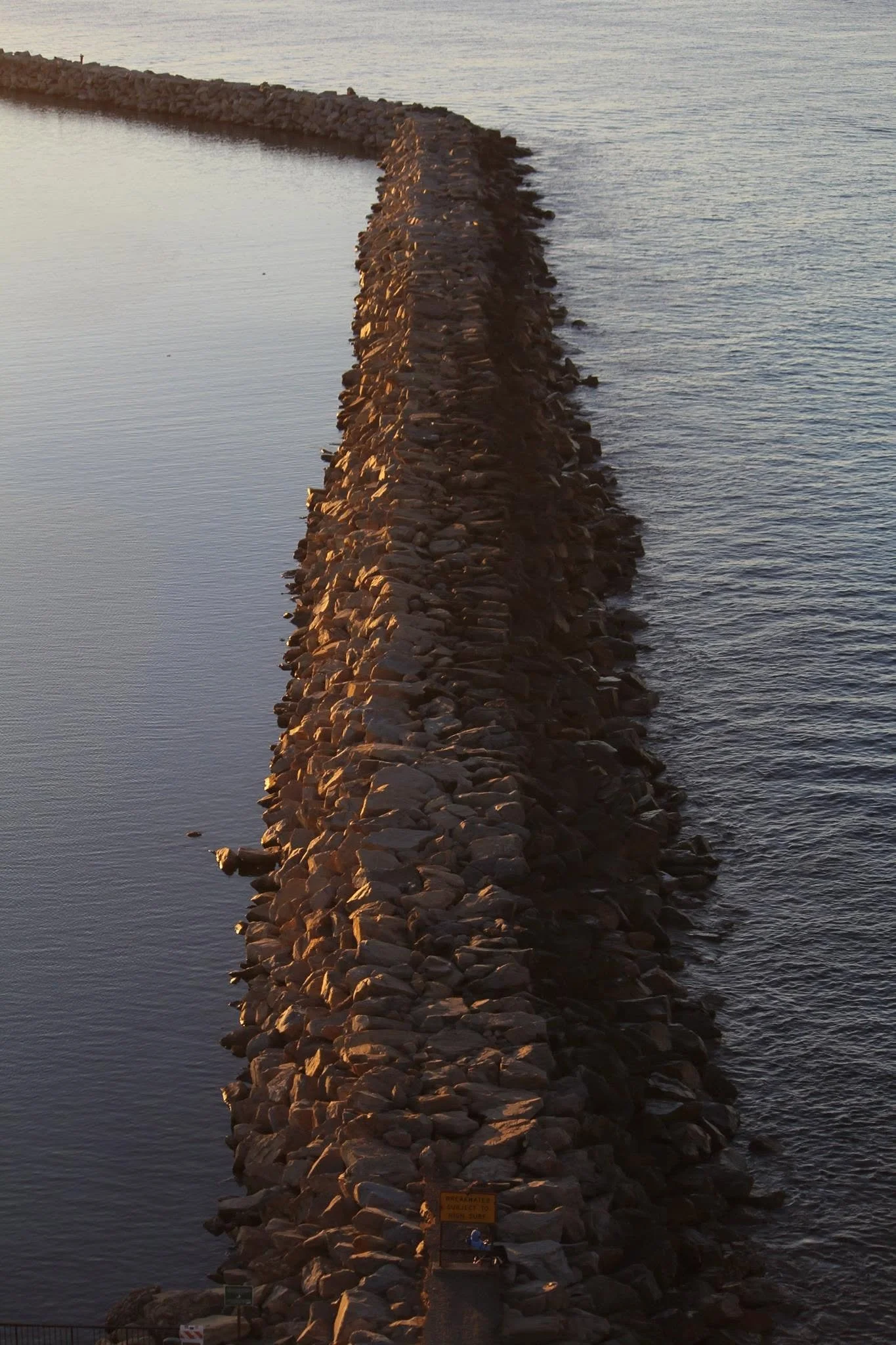 A rocky breakwater extends into the water, with a small sign at the bottom. The water on either side is calm, and the sun casts a warm glow, suggesting late afternoon or early evening.