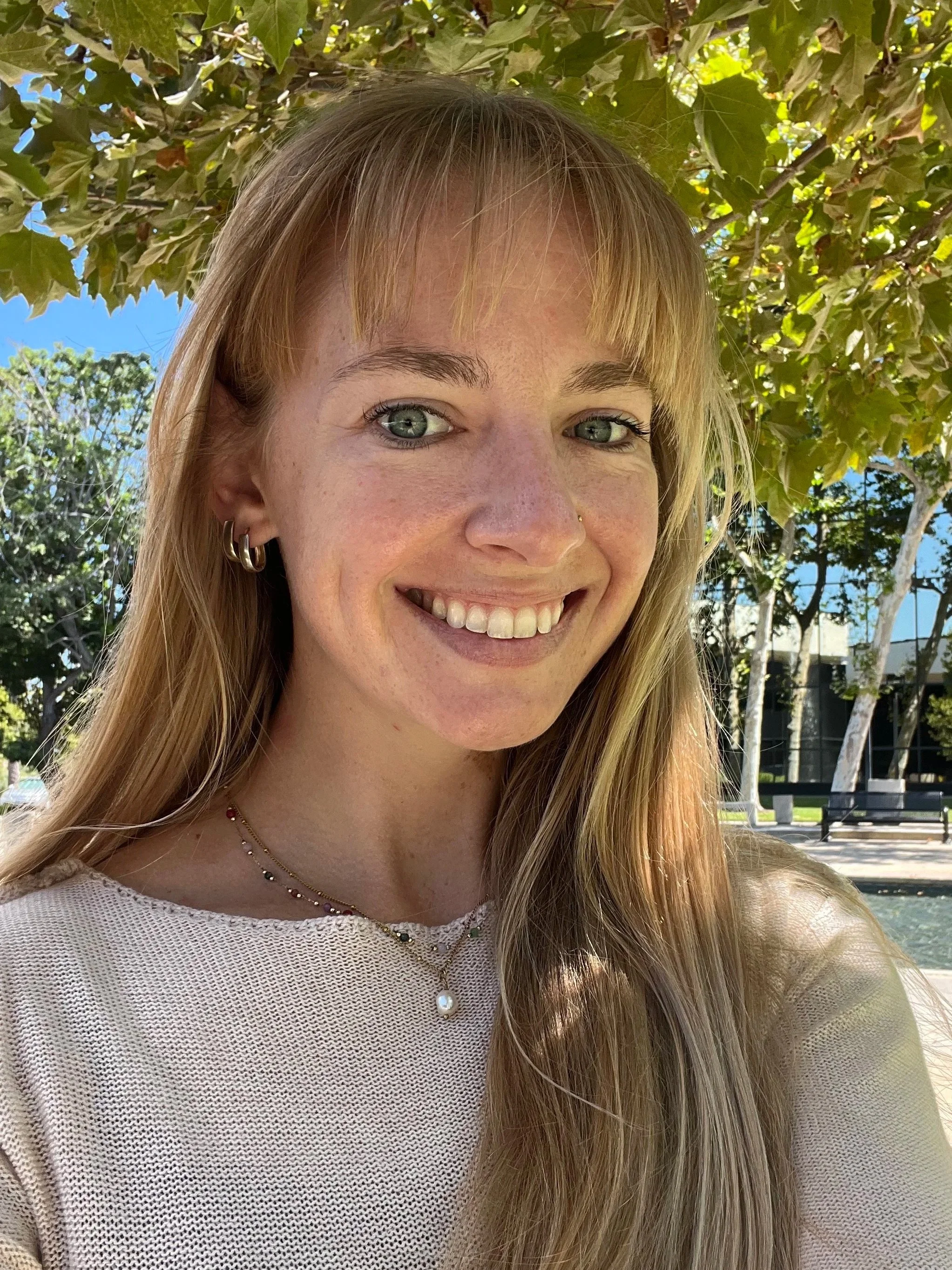 A young woman with blonde hair and green eyes smiling outdoors, wearing earrings and layered necklaces, with trees and a bench visible in the background.
