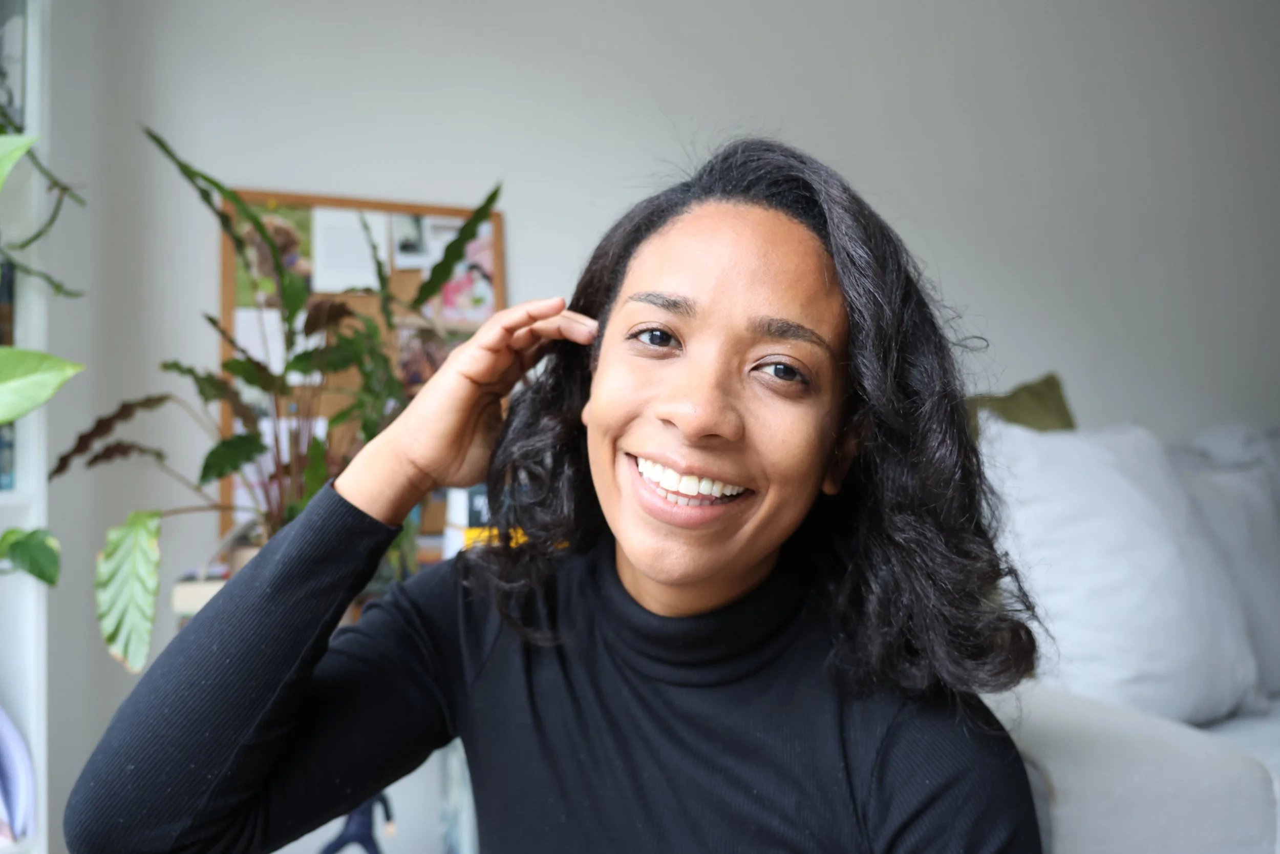 A smiling woman with wavy dark hair in a black top, sitting indoors near several potted plants with green leaves, in a cozy room with white walls and a bulletin board in the background.