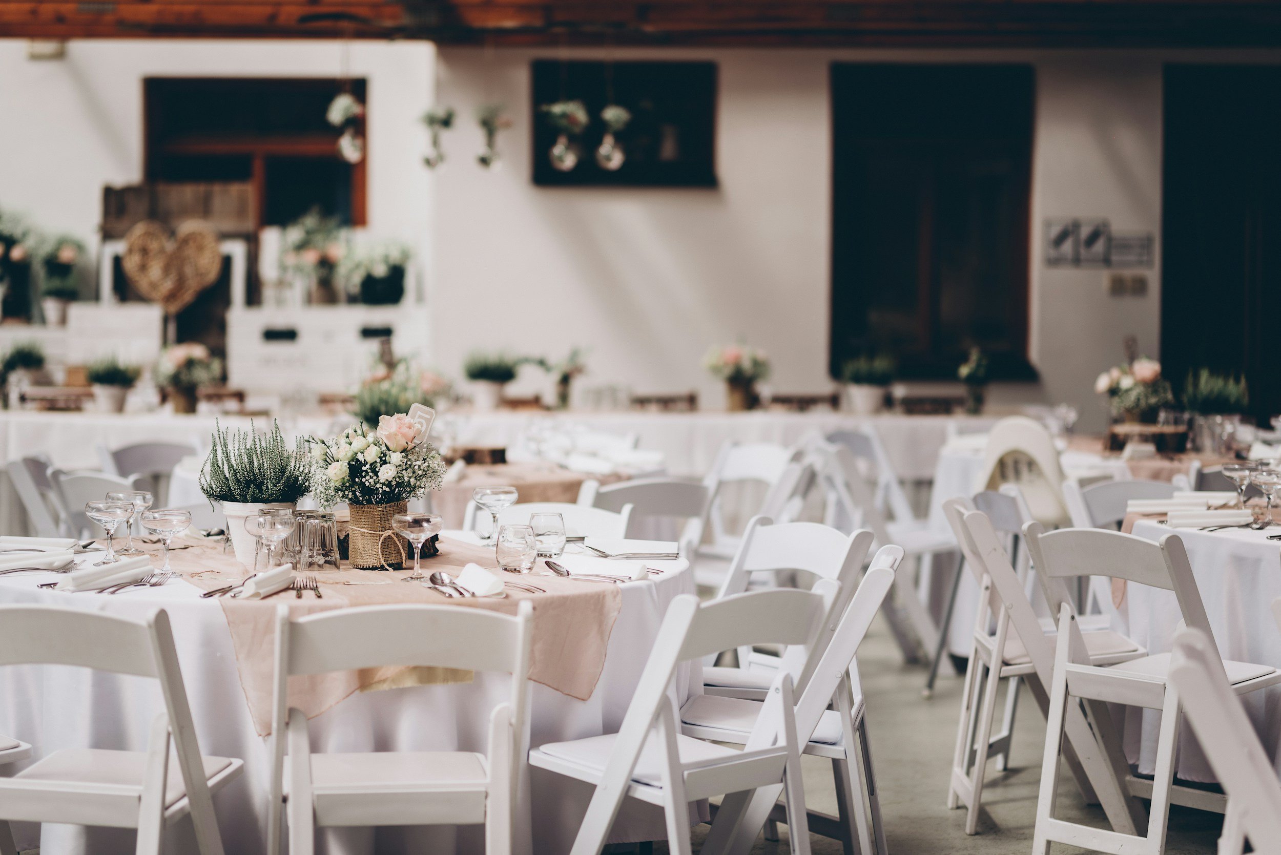 Indoor event space decorated for a celebration with white chairs and tables, floral centerpieces, and a wall with hanging plants and decorations.