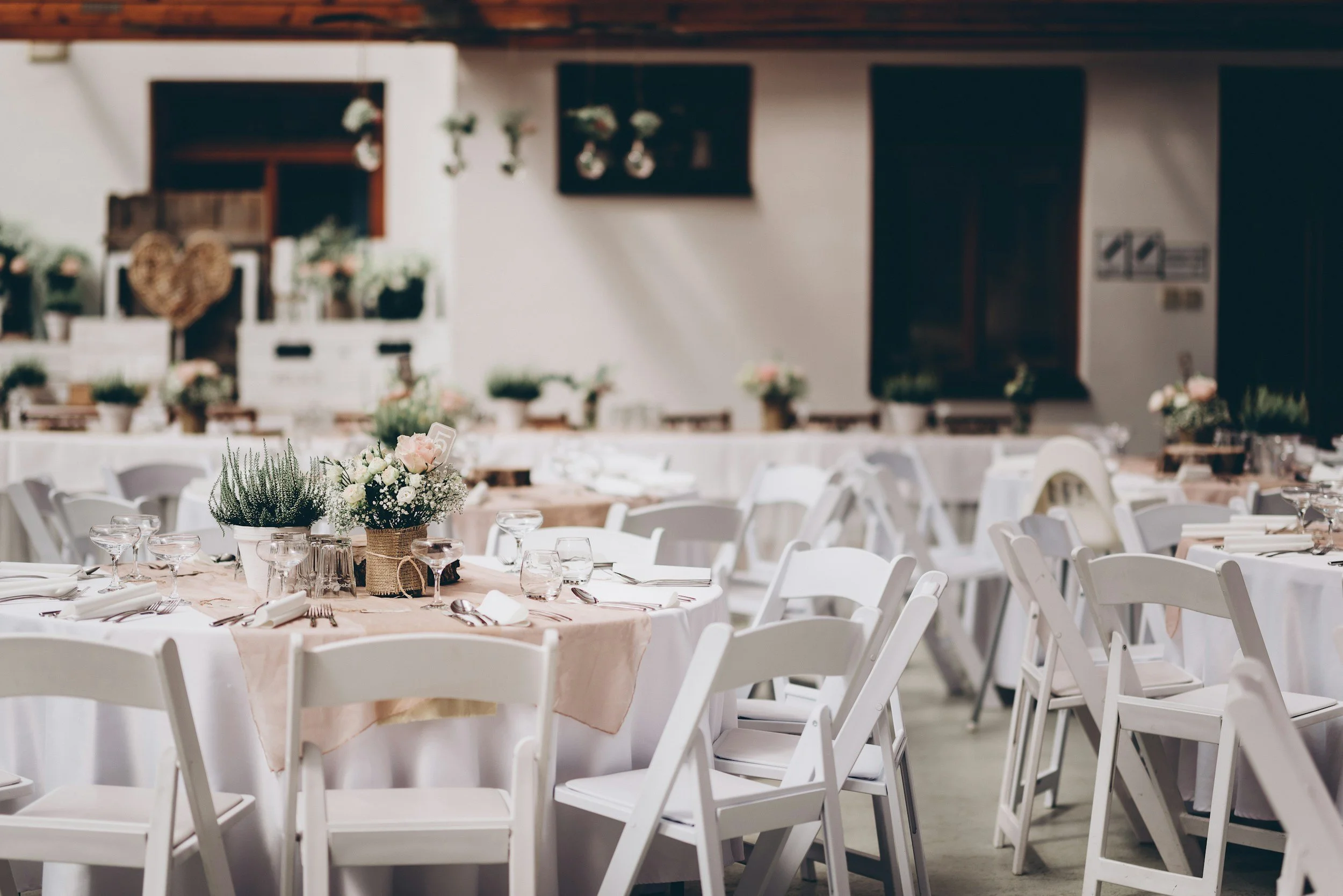 Elegant banquet hall decorated with white chairs, round tables with light pink tablecloths, flower centerpieces, glassware, and silverware, set for a formal event.