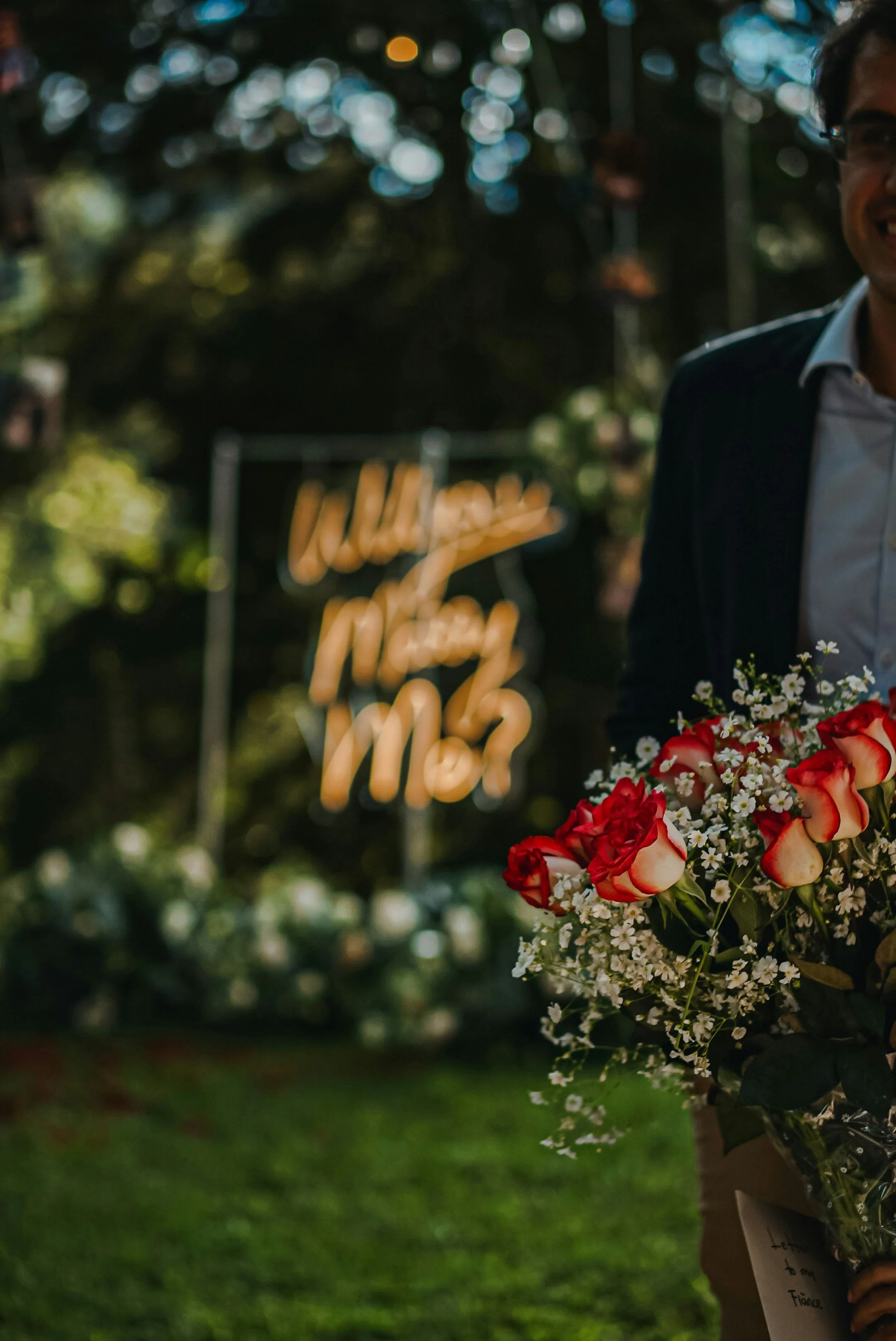 Person holding a bouquet of red and white flowers, standing outdoors with greenery and a blurred sign in the background.
