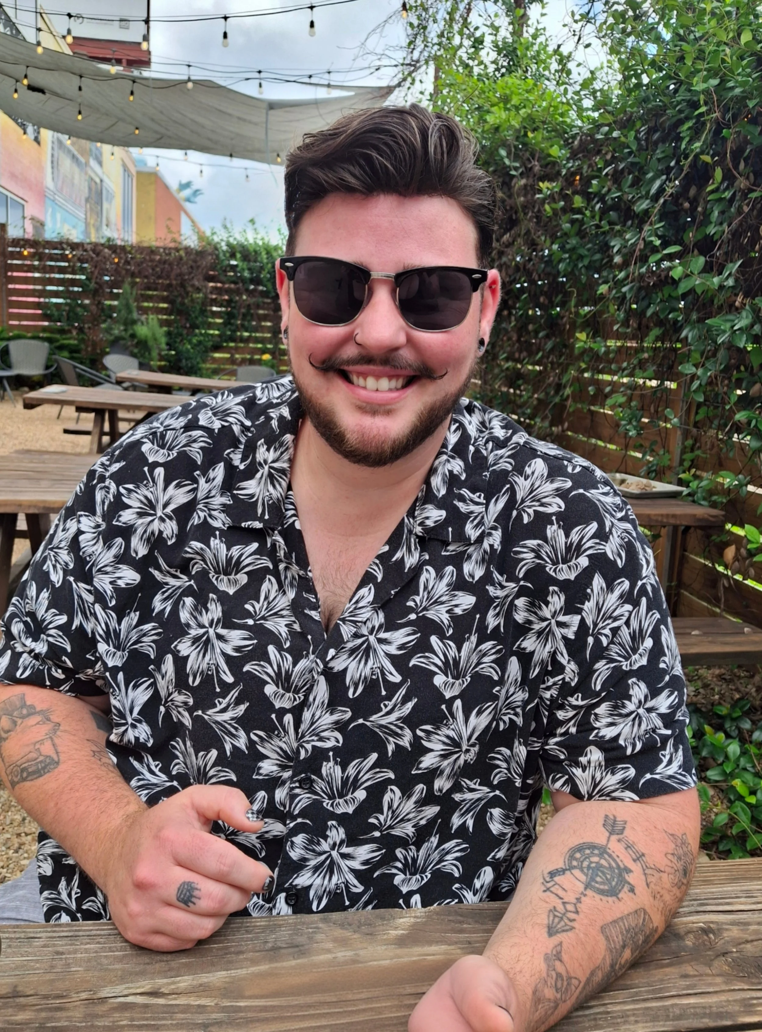 A man with dark sunglasses, a nose ring, a painted mustache, and a floral shirt sitting at a wooden table outdoors, smiling, with tattoos on his arms, in a garden with green foliage and colorful buildings in the background.