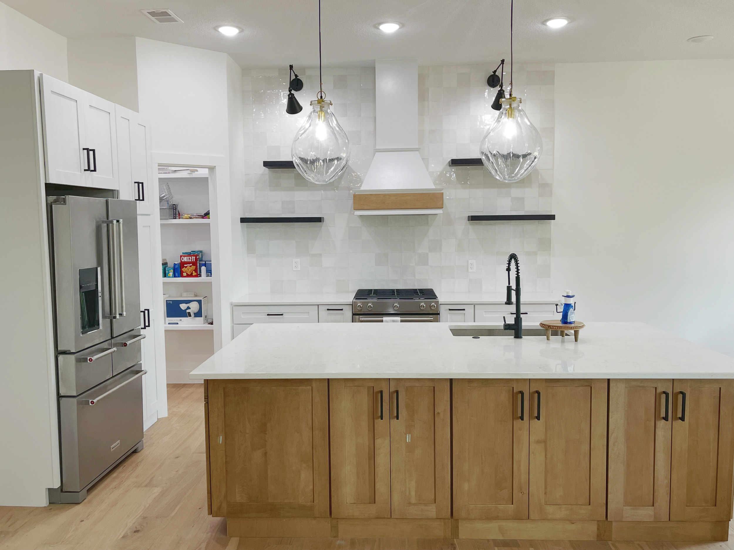 A modern kitchen with a white island countertop, wooden cabinetry, a stainless steel refrigerator, open white shelves, a stove, a white range hood, black floating shelves, a black faucet, and two large pendant lights with clear glass globes hanging from the ceiling.