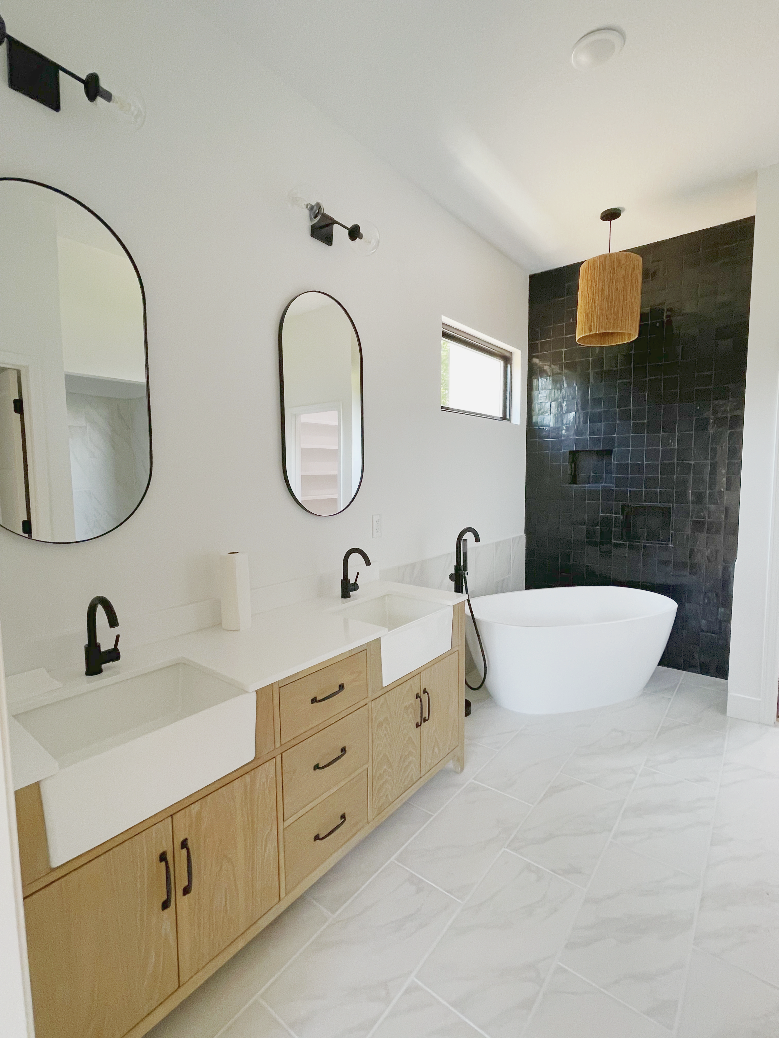 Modern bathroom with a white freestanding bathtub, a double vanity with black faucets, and a black tiled wall with a built-in niche and a wooden pendant light hanging from the ceiling.