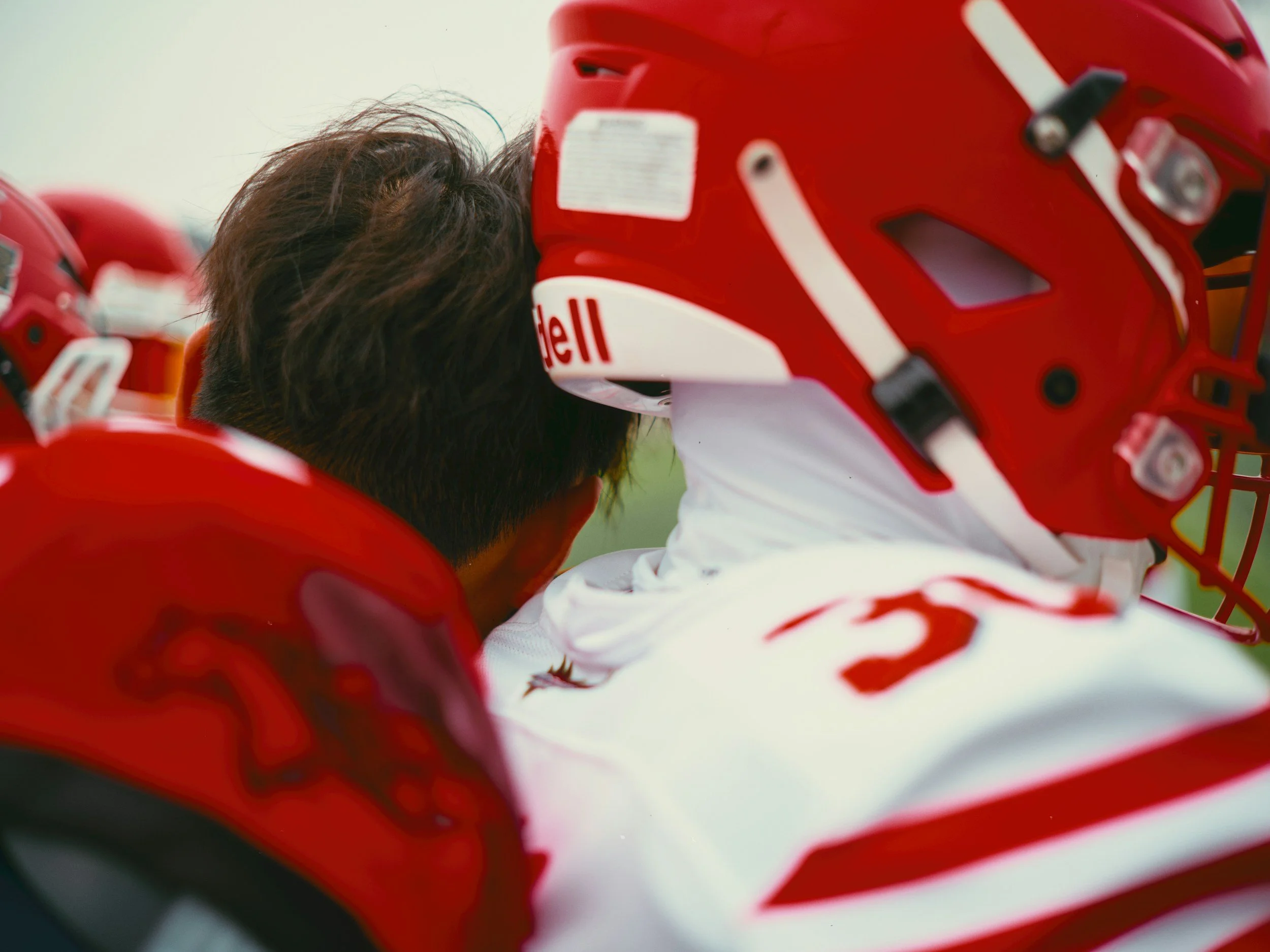 Close-up of a person wearing a red football helmet with white accents, leaning forward with dark hair visible, with a second similar helmet partially visible in the foreground.