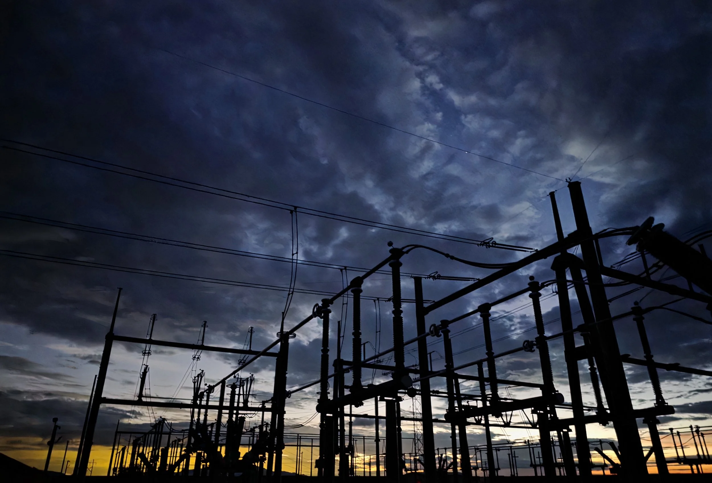 Power lines and electrical infrastructure silhouetted against a sunset sky with dark clouds.
