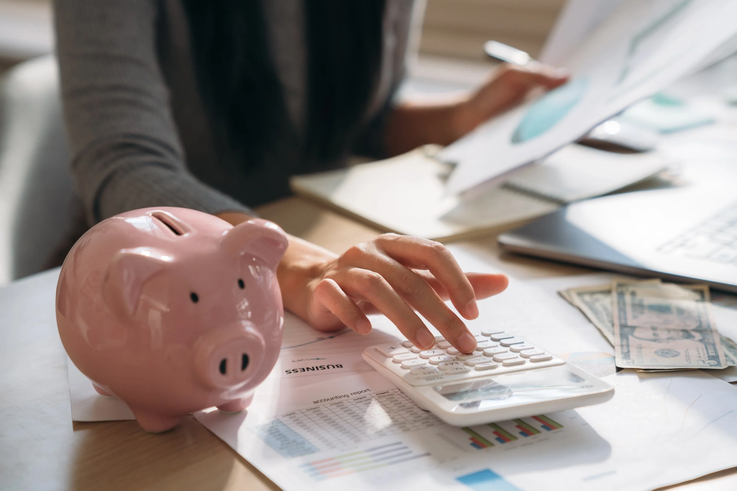 A pink piggy bank, a person using a calculator, and money on a table with documents and charts.