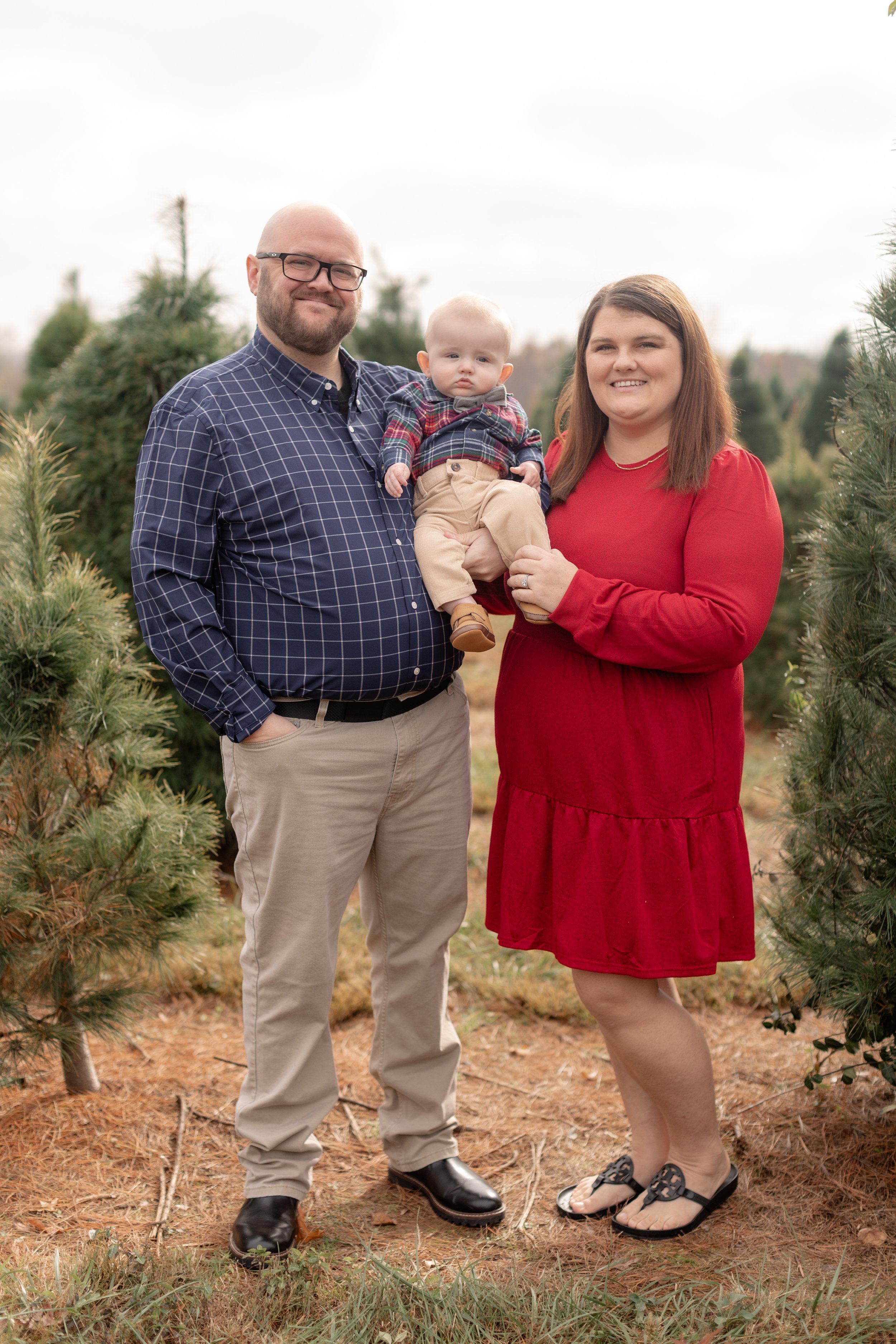 A family of three posing outdoors among evergreen trees. The man with glasses, beard, and checkered shirt is standing on the left, holding a baby who is dressed in plaid shirt and beige pants. The woman in a red dress is standing on the right, smiling at the camera and holding the baby.