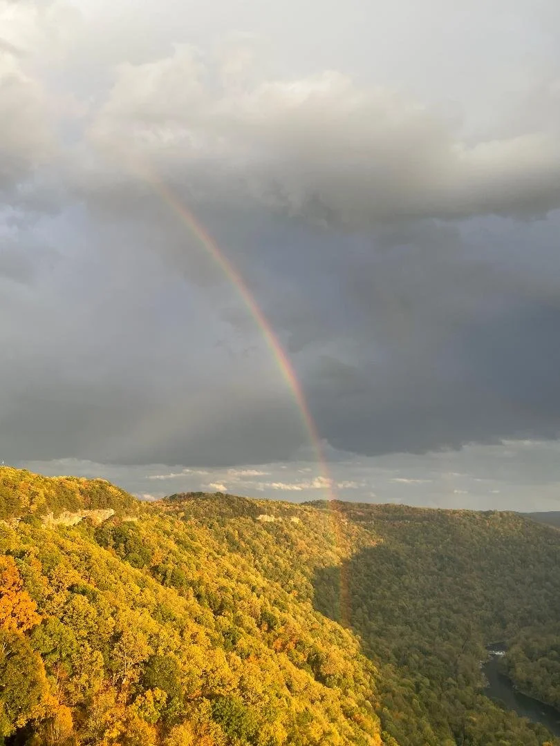 Rainbow arching over a hilly landscape with dense trees showing fall colors and a river at the bottom.