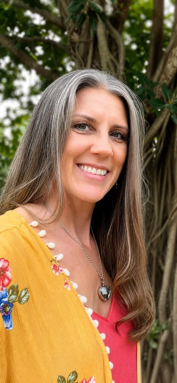 A smiling woman with long gray hair outdoors, wearing a red top and a yellow shawl with floral embroidery, jewelry including a heart-shaped locket necklace and pearl accents, standing in front of a tree with green foliage.
