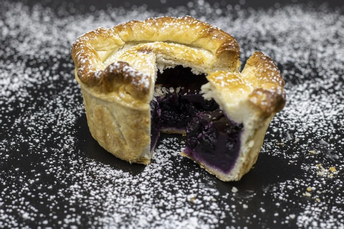 An open blueberry cream pie with a golden crust, dusted with powdered sugar, on a black surface sprinkled with powdered sugar.