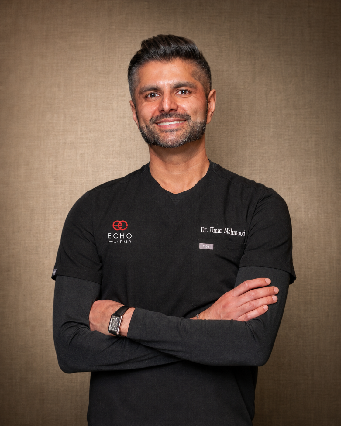 A man with dark hair and a beard wearing a black medical scrubs top with a logo and the name 'Dr. Umar Mahmood' on it, standing with arms crossed against a beige wall.