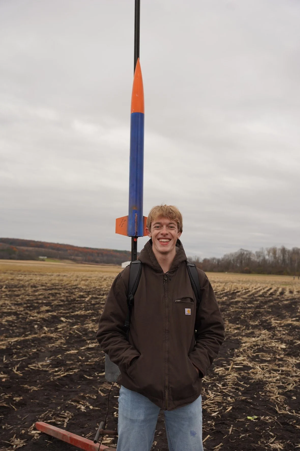 A young man with brown hair, wearing a brown Carhartt jacket and light blue jeans, smiling and standing outdoors in a plowed field under an overcast sky. Behind him, there is a tall, vertical model rocket with an orange nose cone, blue body, and orange fins, mounted on a launch pad.