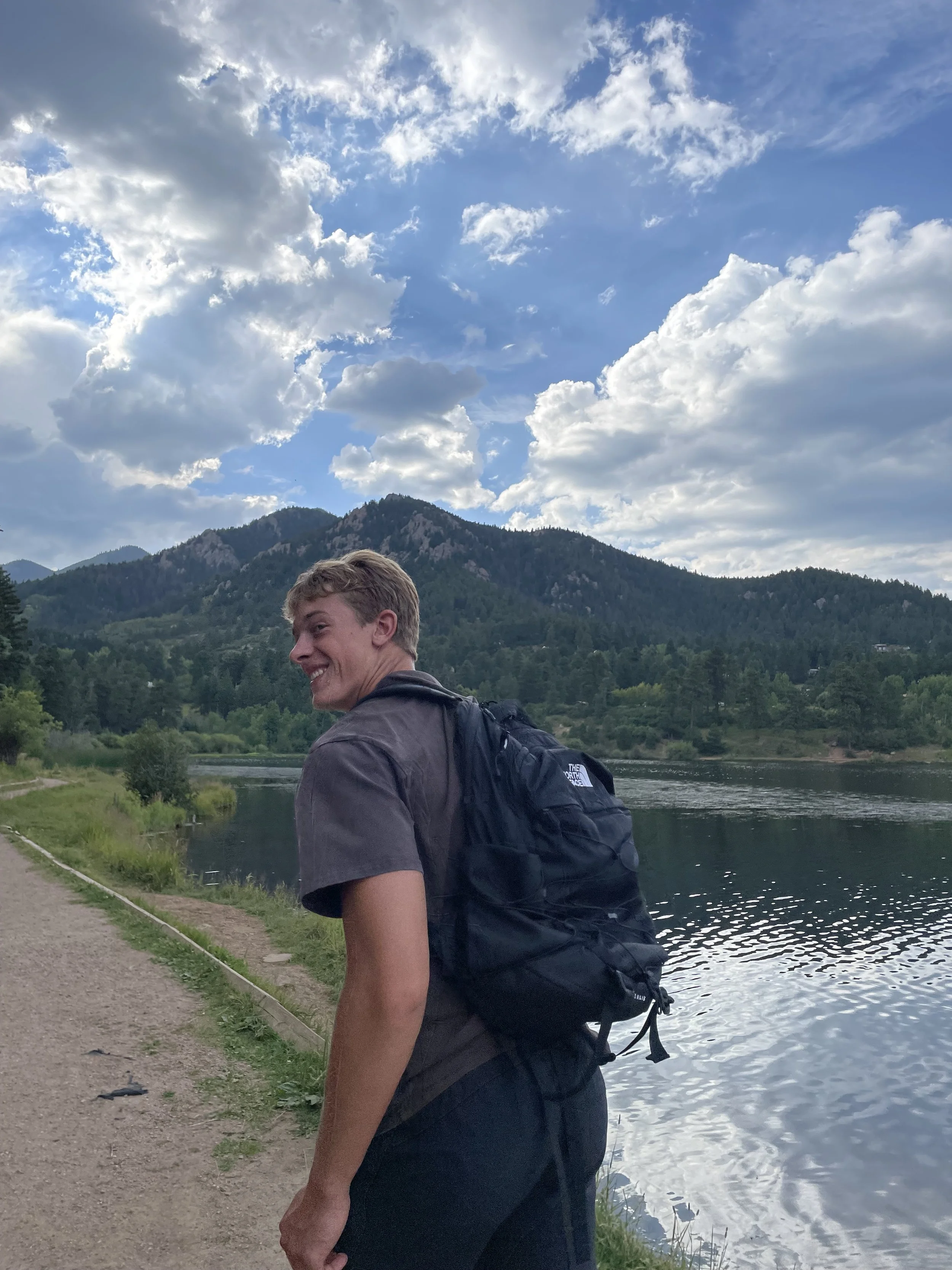 Young man with short blond hair smiling, wearing a dark t-shirt and backpack, standing on a riverside trail with mountains and a cloudy sky in the background.