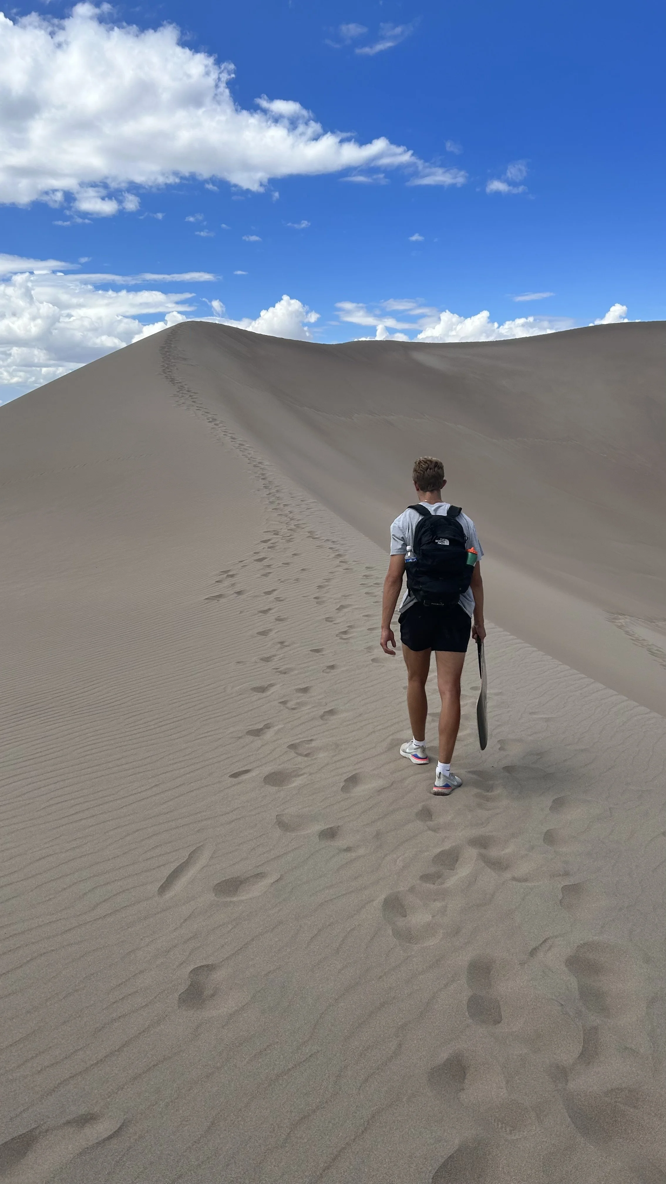 Person with a backpack walking on a sandy desert dune with footprints, under a blue sky with clouds.