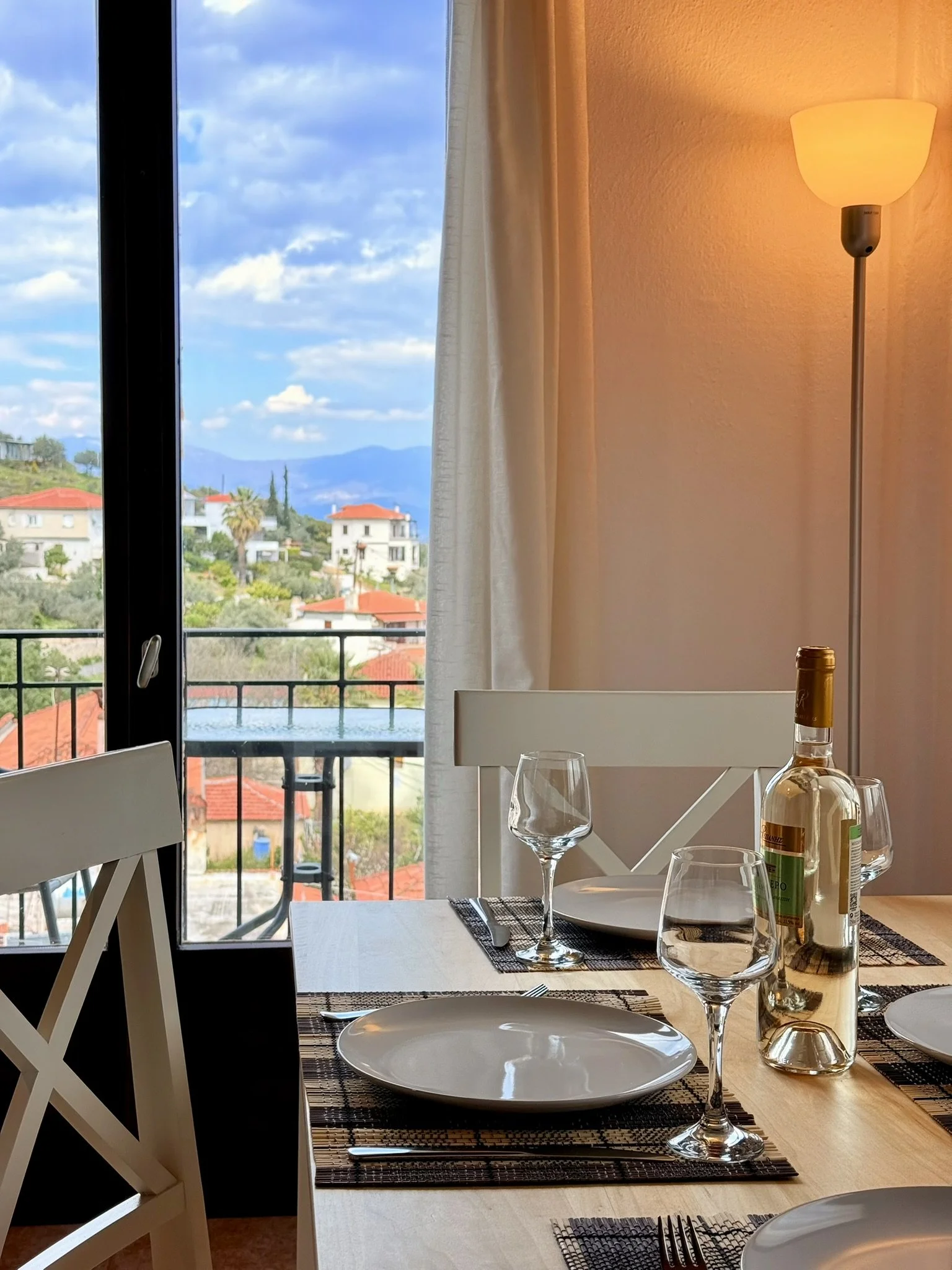 Interior of a dining area with a table set for a meal, featuring white plates, wine glasses, a bottle of wine, and silverware, with a large window showing a landscape of houses, trees, and mountains in the background, and a warm-colored wall with a l