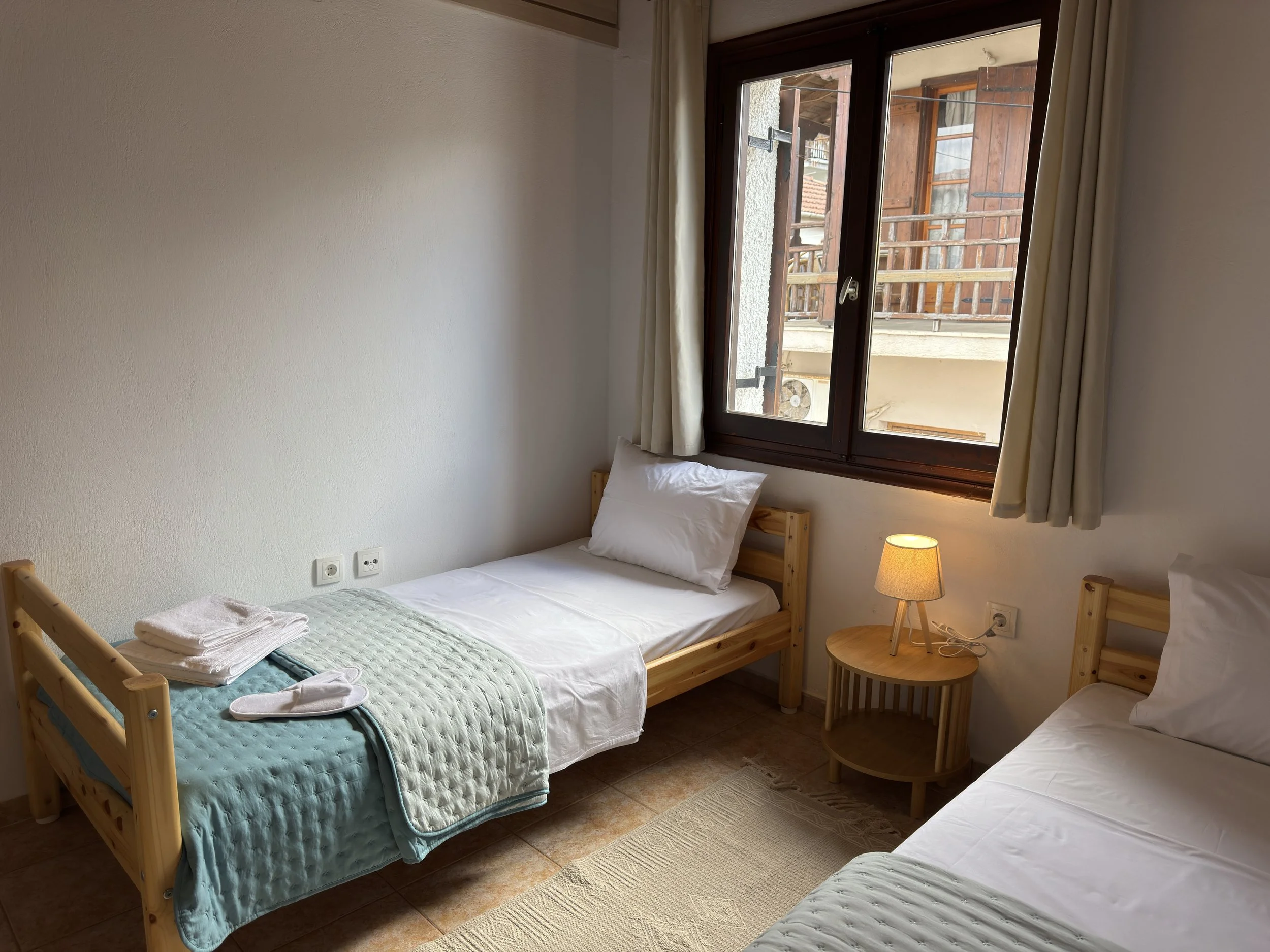 A bedroom with two beds, a window with curtains, a small round side table with a lamp, and towels on one of the beds.