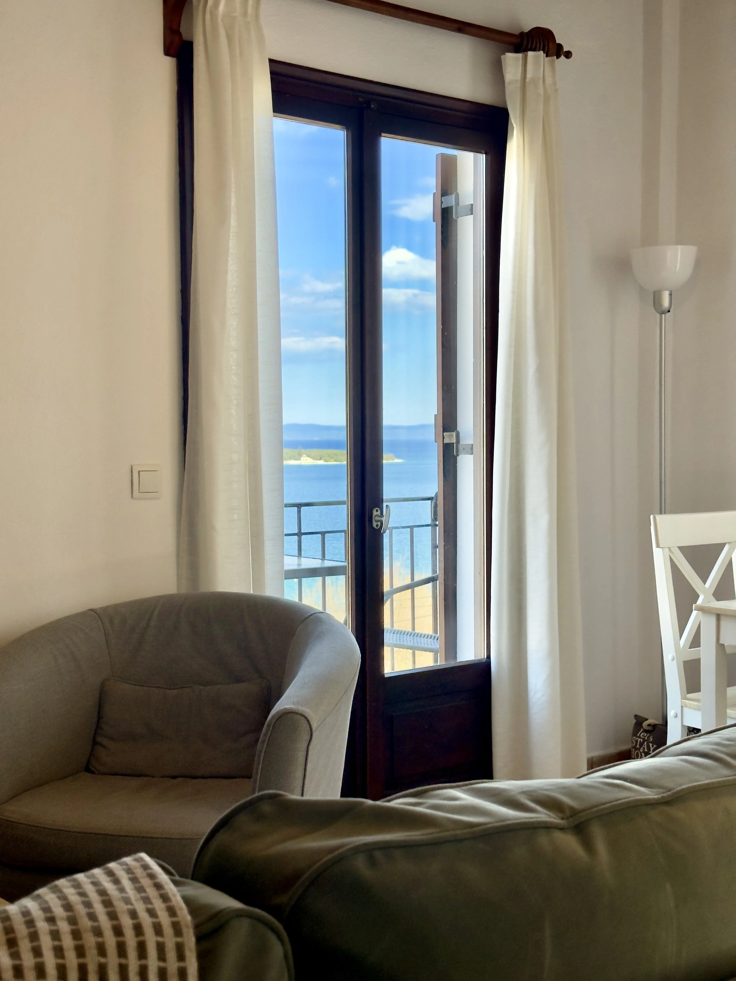 Living room with large open door leading to balcony and scenic ocean view, beige armchair, white curtains, and floor lamp.