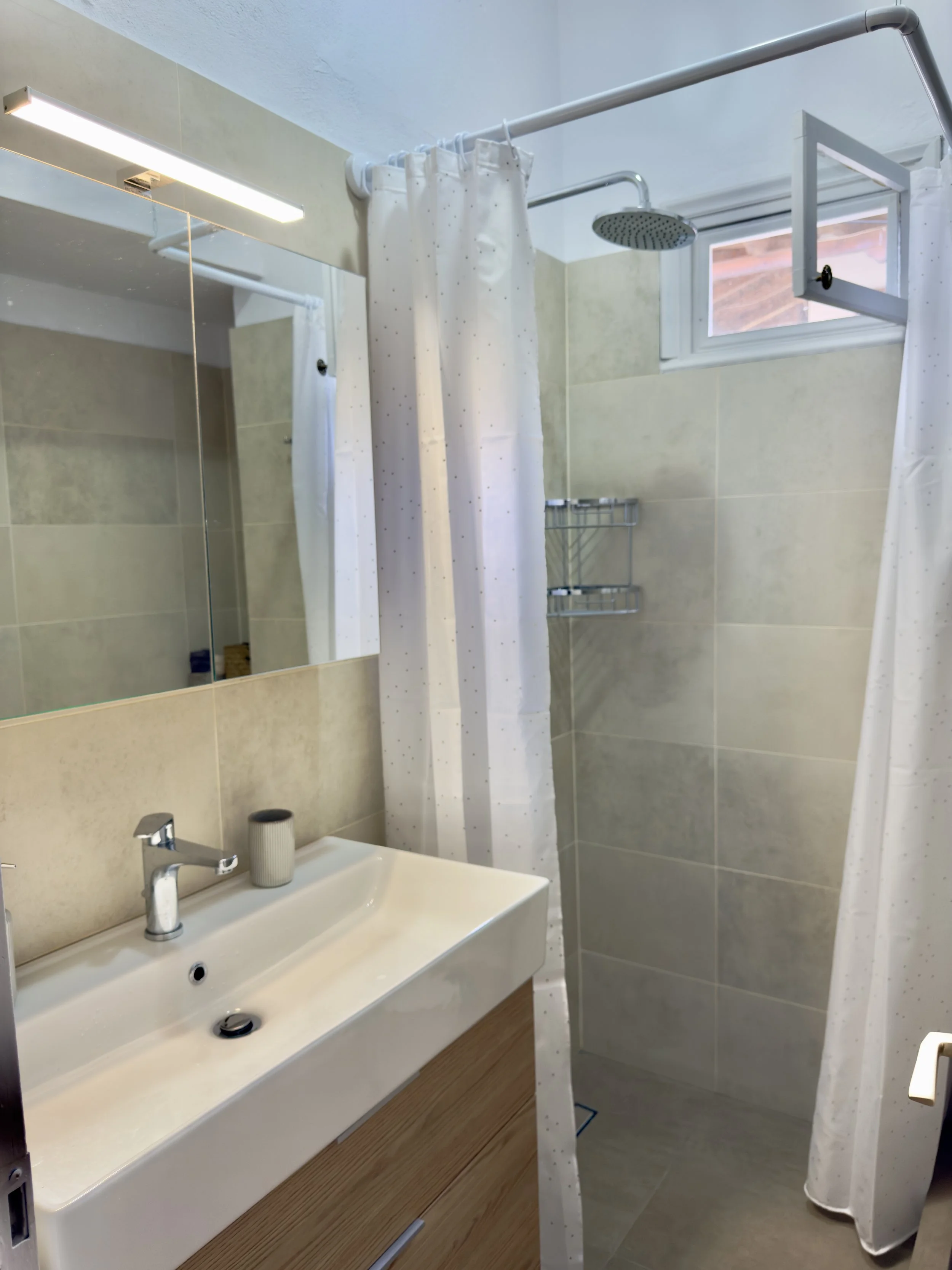 Bathroom with a rectangular white sink, a faucet, a small gray cup, a mirror above the sink, a shower with a white curtain, a window with a view outside, beige tiled walls, and a wooden vanity below the sink.