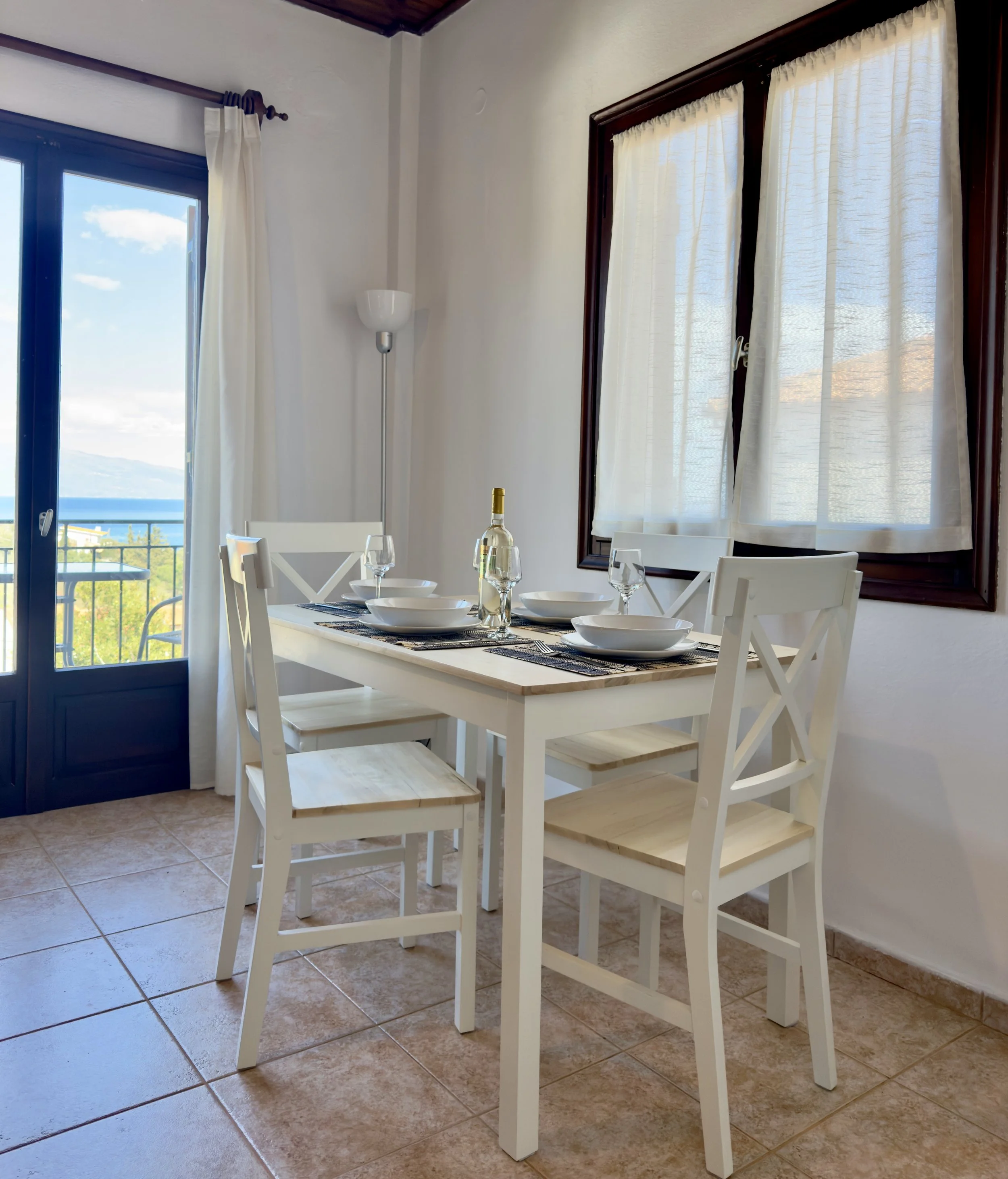 A dining area with a white wooden table set with white bowls, glasses, and a bottle of wine, surrounded by white chairs, and large windows with light curtains revealing a scenic outdoor view.
