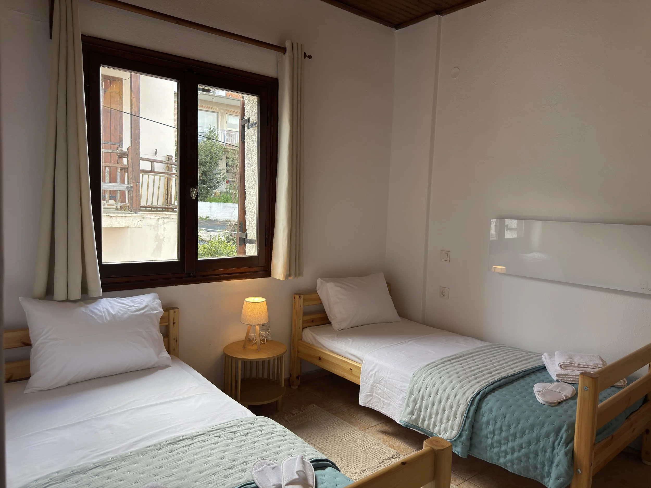 A bedroom with three single beds made of light wood. Two beds are on the left, and one on the right. Each bed has white bedding and pillows. The bed on the right has a greenish quilt and a pair of slippers on it. A small round wooden nightstand with a lamp is positioned between the two beds on the left. The wall has a window with beige curtains. Outside the window, there are buildings and greenery.