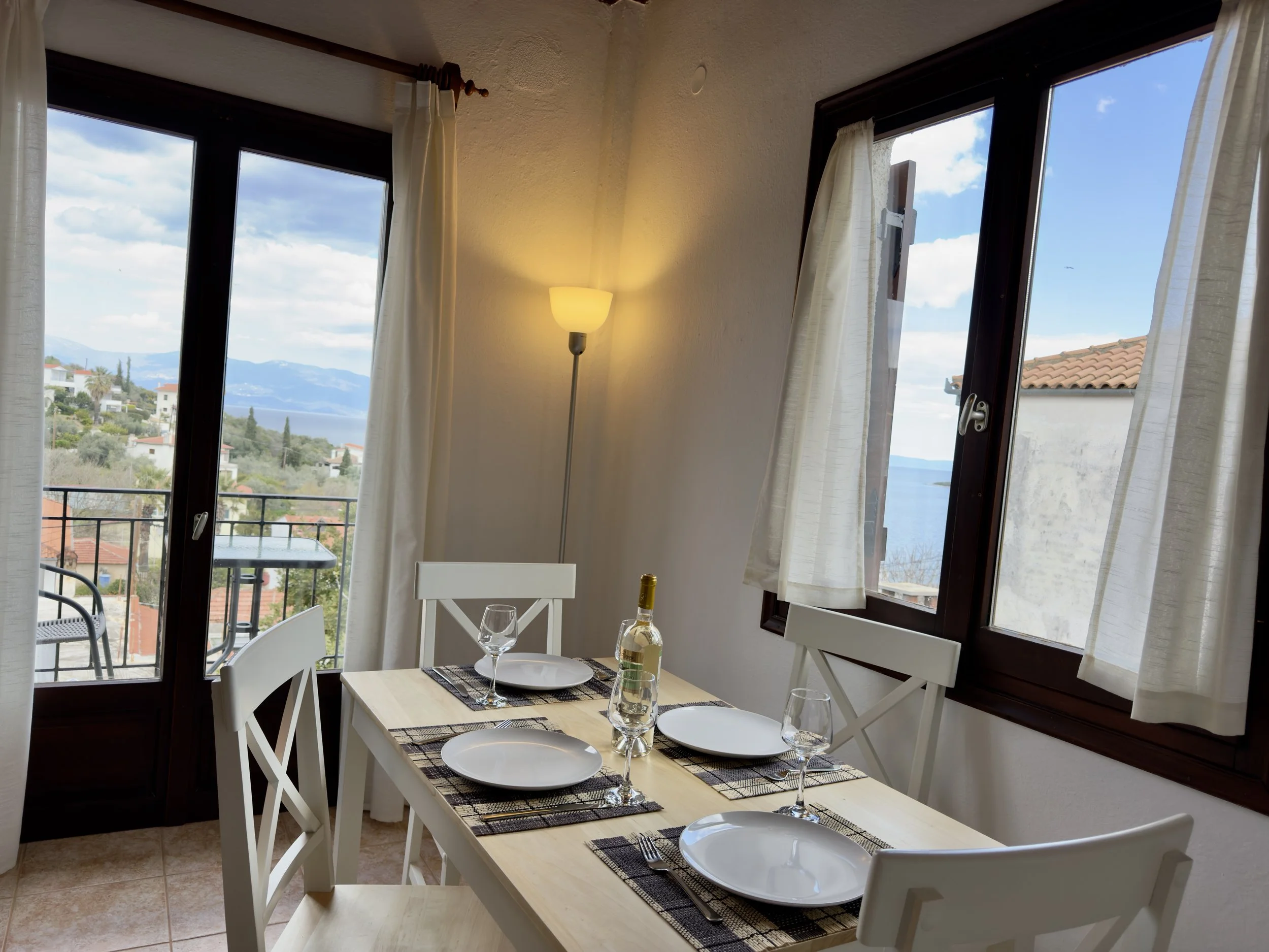 Dining table set for four with white plates, wine glasses, a bottle of wine, and silverware in a room with large windows showing a scenic view of mountains, blue sky, and a lake.