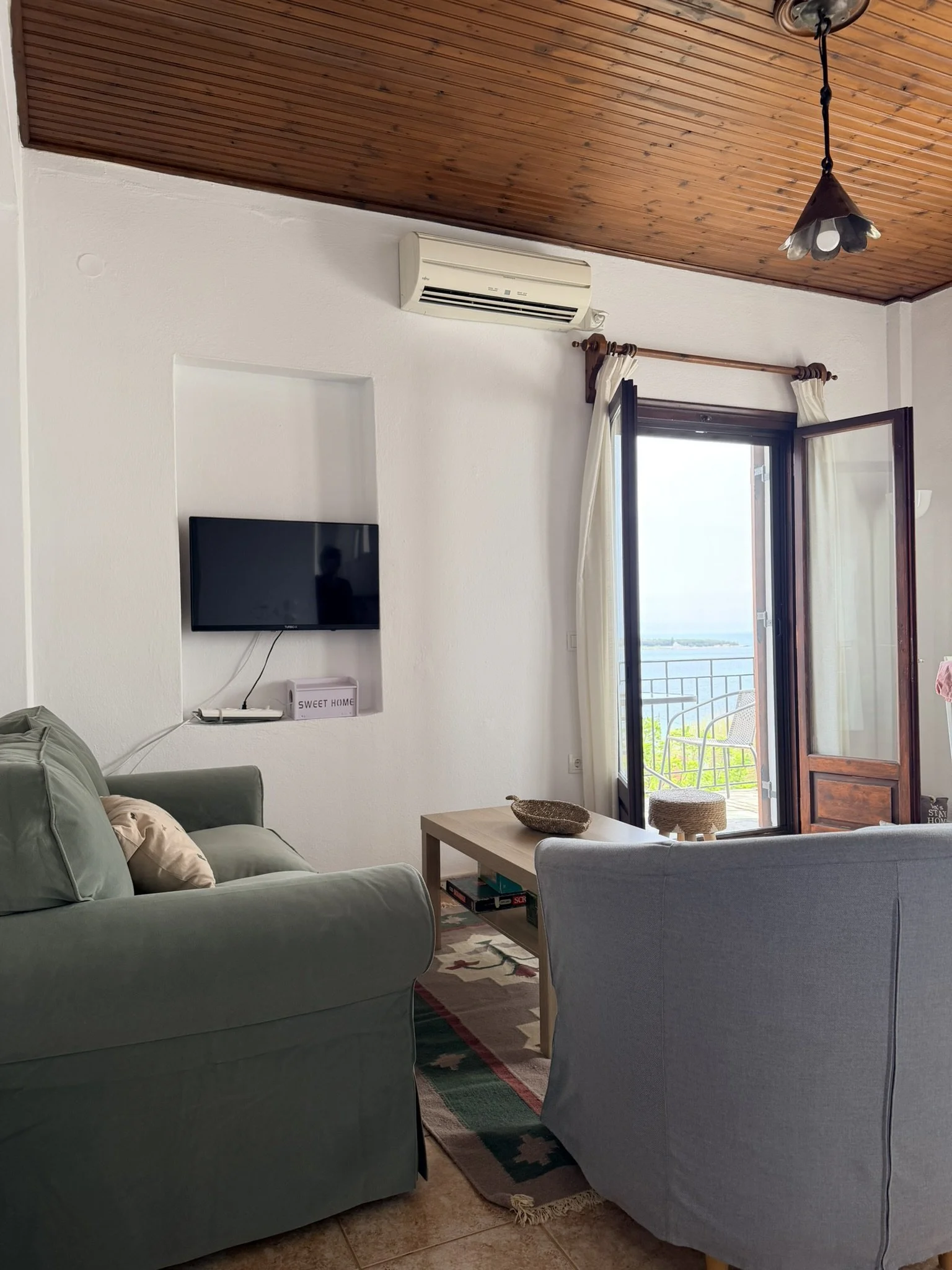 A cozy living room with a green sofa, a wooden coffee table, a wall-mounted flat-screen TV, and a window with white curtains showing an outdoor balcony with a view of the water.