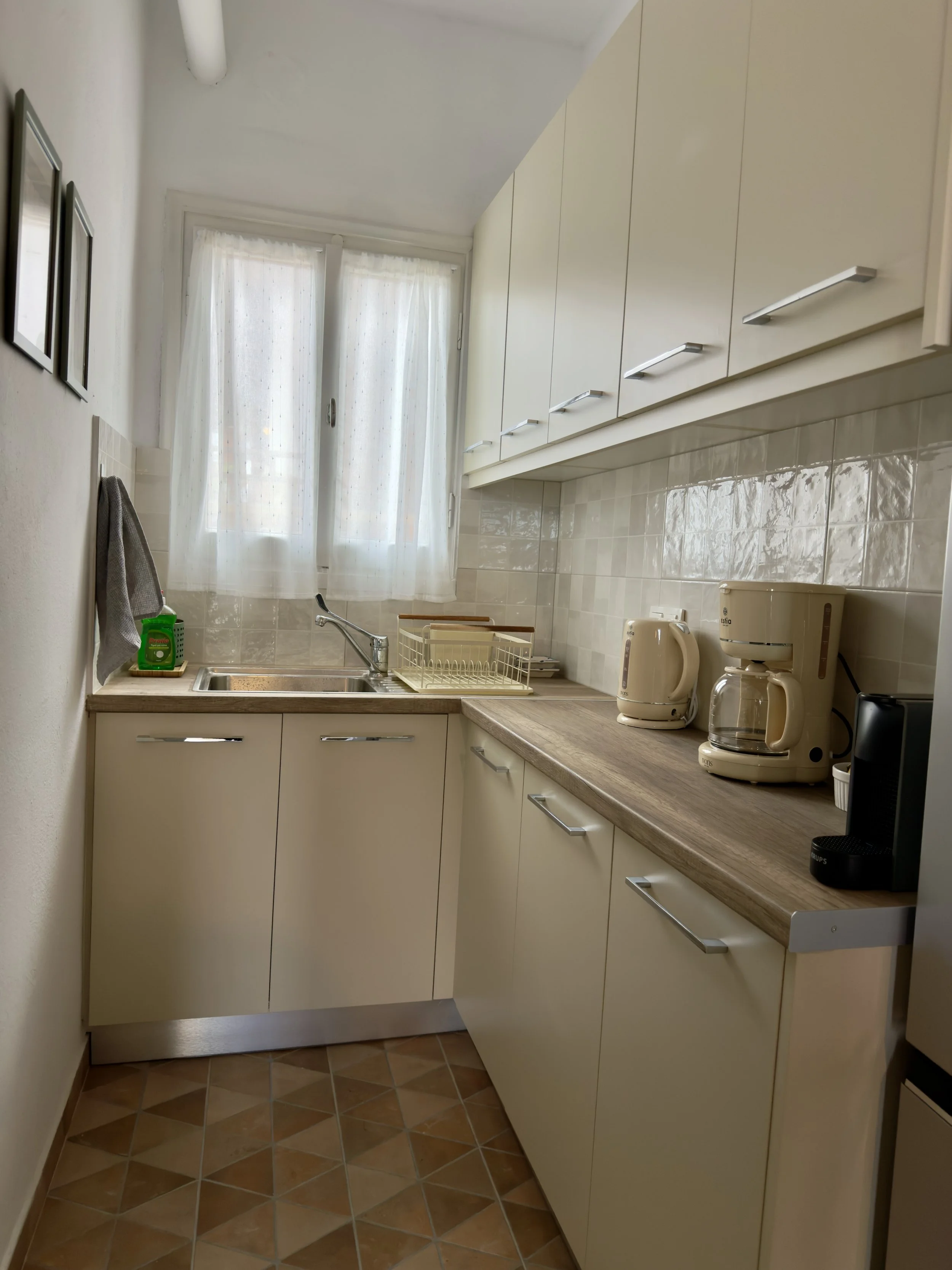Small kitchen area with white cabinets, a window with curtains, a sink, a dish drying rack, a coffee maker, and a kettle on a wooden countertop.