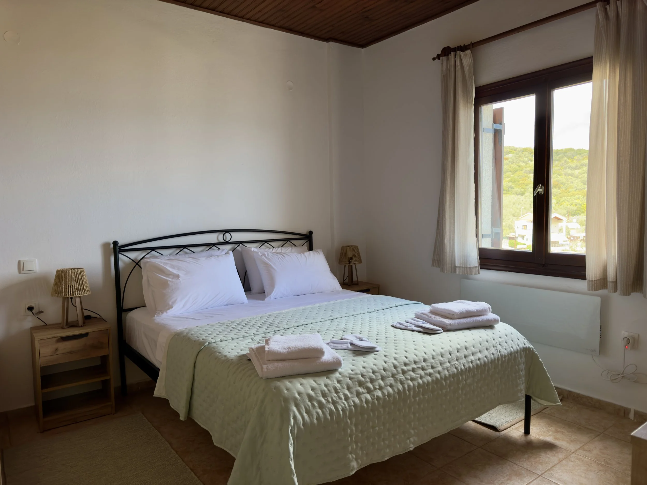 A bedroom with a bed that has a green quilt, white pillows, and towels on top. Two wooden nightstands with wicker lamps flank the bed. There is a large window with beige curtains, showing a view of trees and buildings outside.