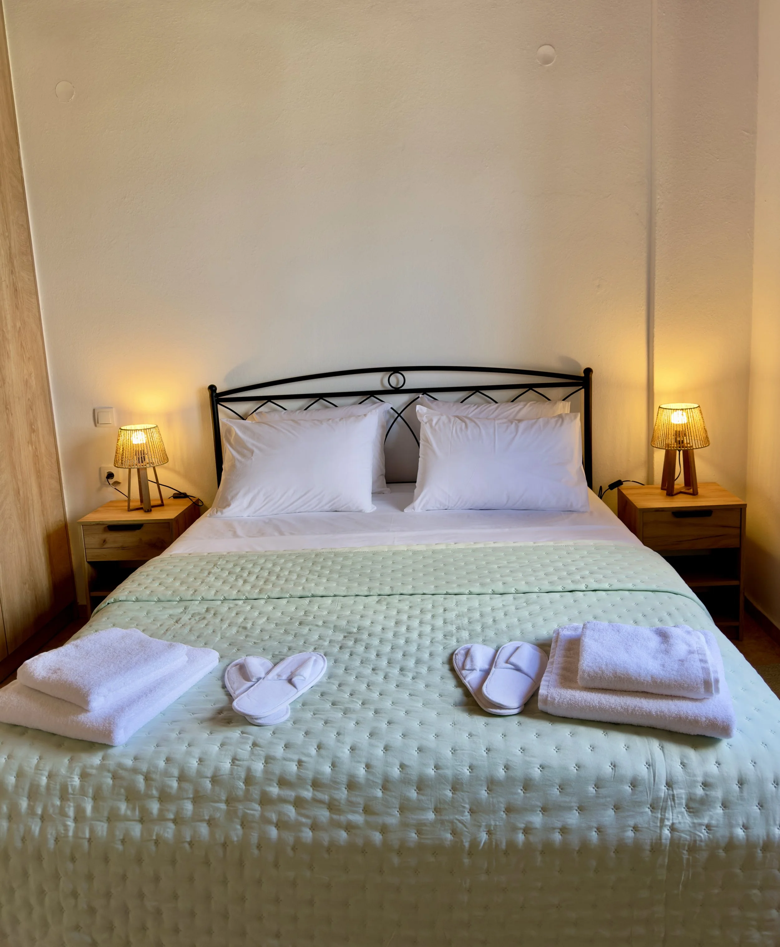 A neatly made bed with a light green quilt, two white pillows, and two rolled white towels at the foot. Two bedside tables with warm yellow lights on either side, and a black metal headboard. The room has a beige wall and wooden flooring.