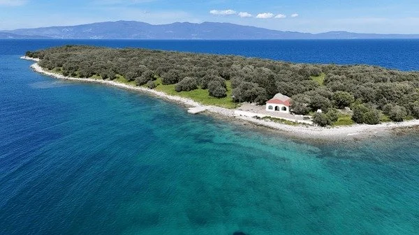 Aerial view of a cove with clear turquoise water, a wooded island, and a small white building with a red roof near the shoreline.