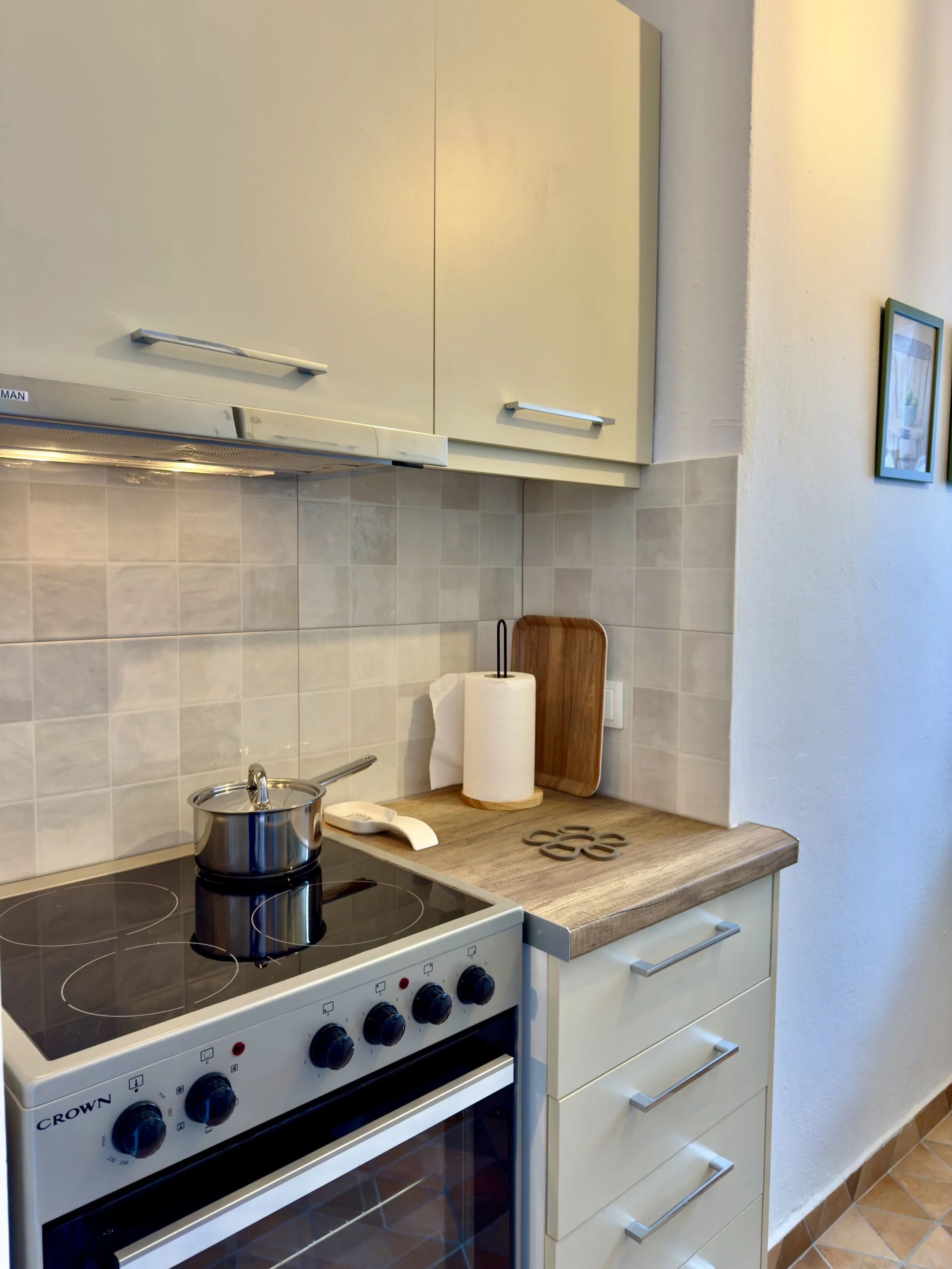 A kitchen counter with a stainless steel pot on an electric stove, a roll of paper towels on a wooden cutting board, a white bowl, and a metal trivet. Light-colored cabinets and tiled backsplash are visible.
