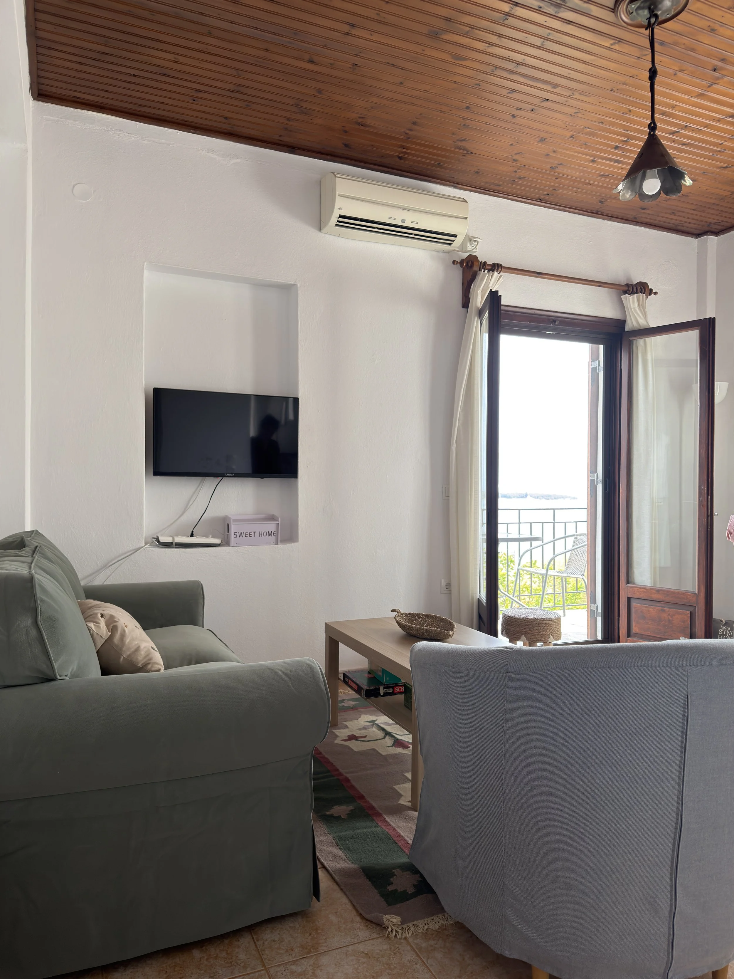 Living room with gray sofa, small wooden table with a stone bowl and a puzzle box, flat-screen TV on white wall, open door leading to balcony with metal chairs, window with cream curtains, wooden ceiling, and a hanging light fixture.