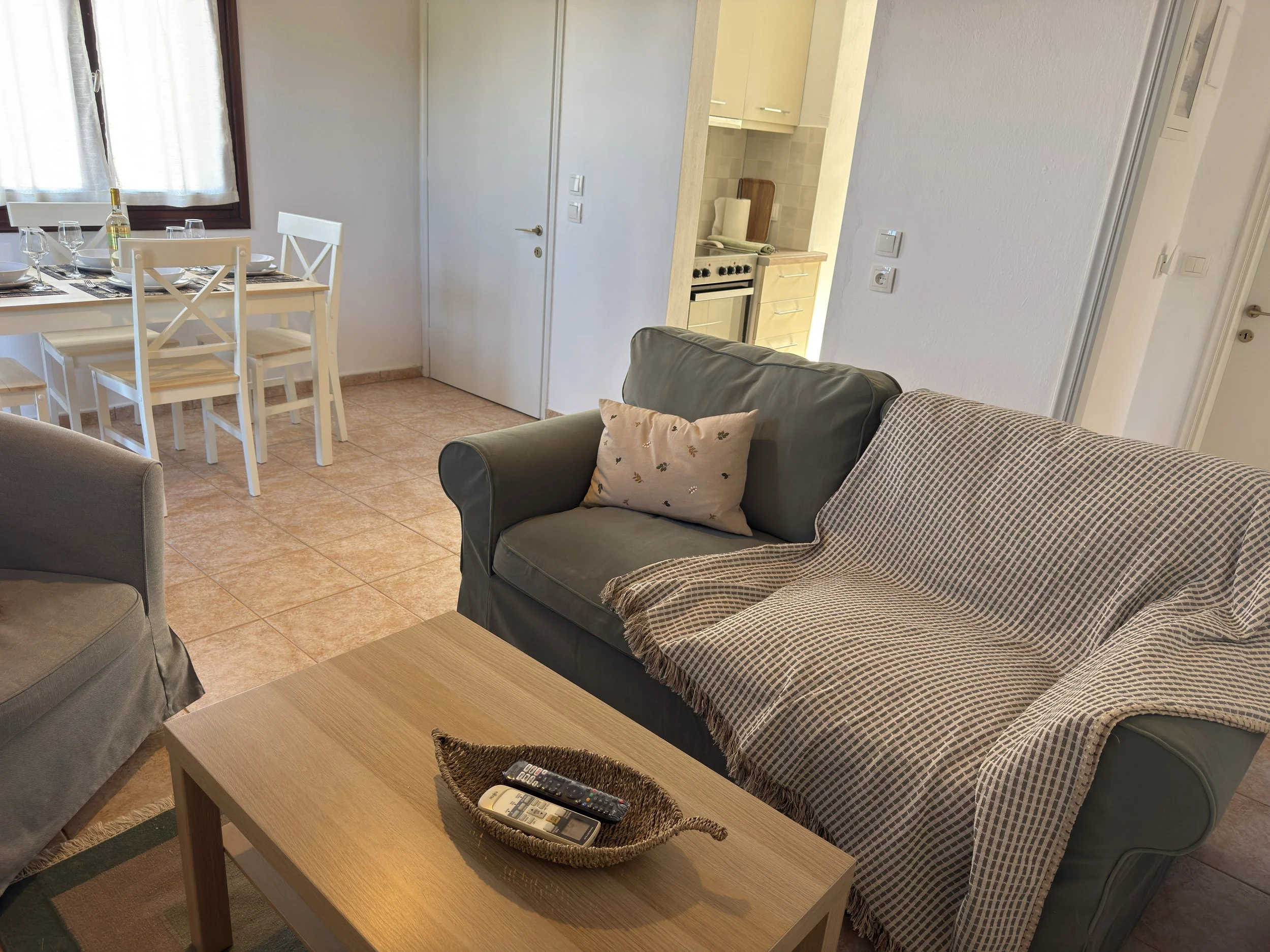 Living room with a wooden coffee table, a basket with remote controls, a sectional sofa with pillows, and a dining area with a table and chairs near a window. Kitchen partially visible.