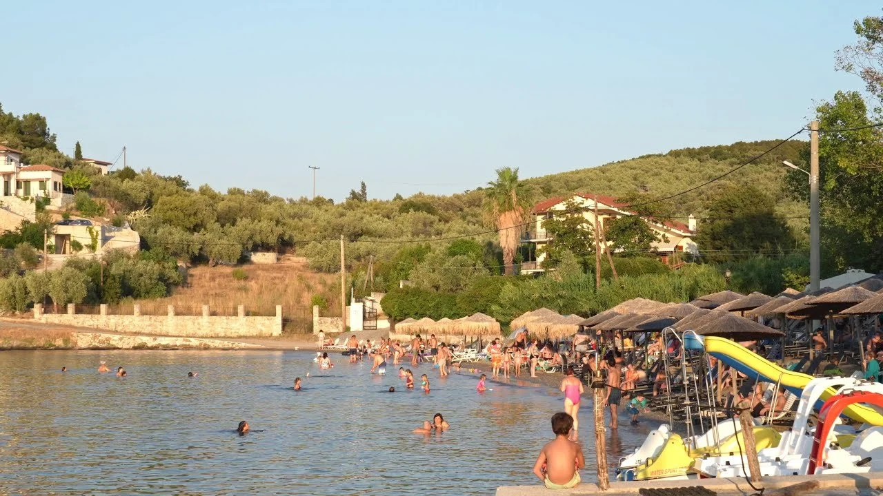Beach with many people swimming and relaxing under umbrellas, with water sports equipment, sand, trees, and hills in the background.