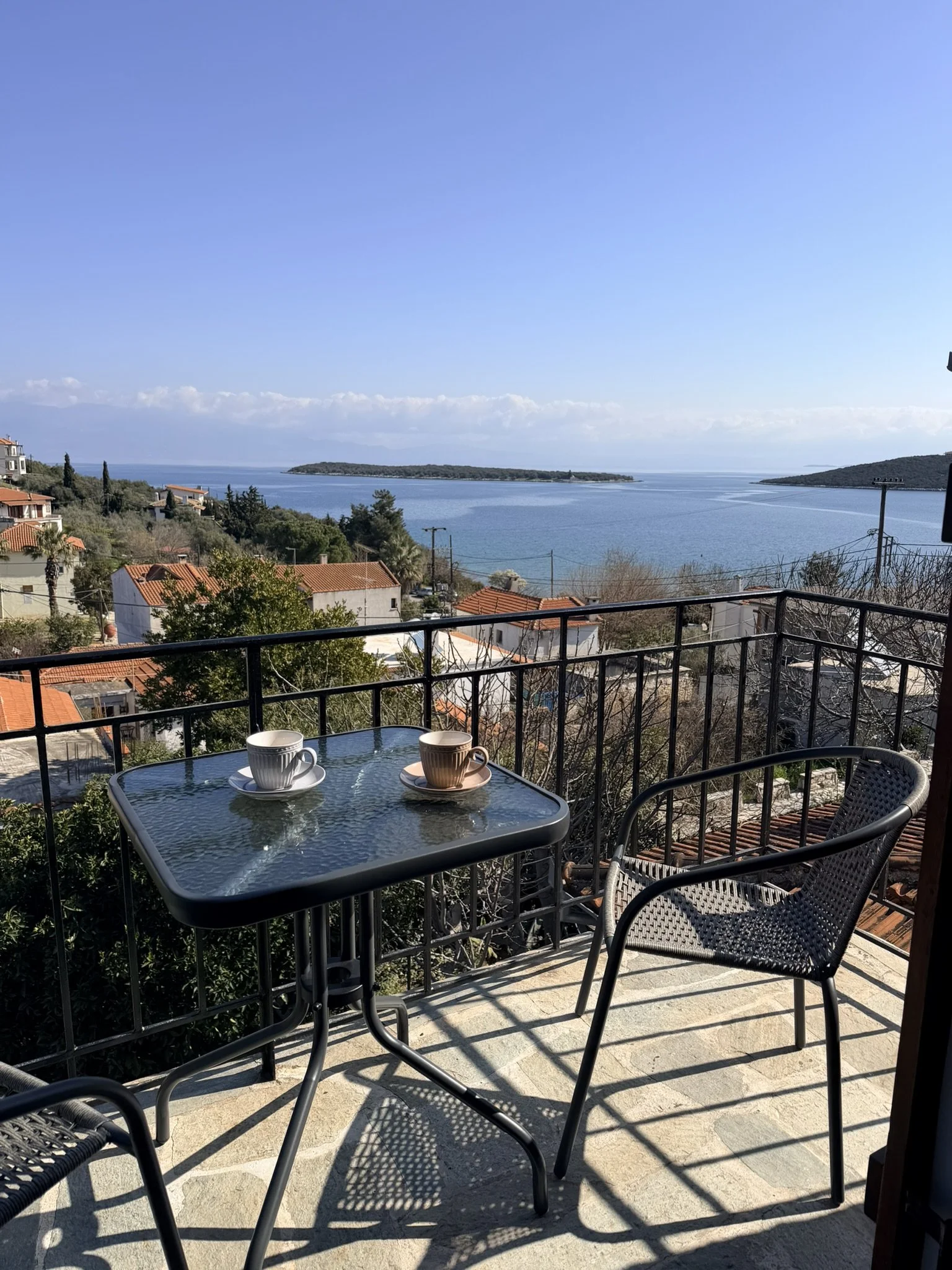 Outdoor balcony with glass table, two cups, and chairs, overlooking a scenic view of the ocean, islands, and houses with red-tiled roofs.