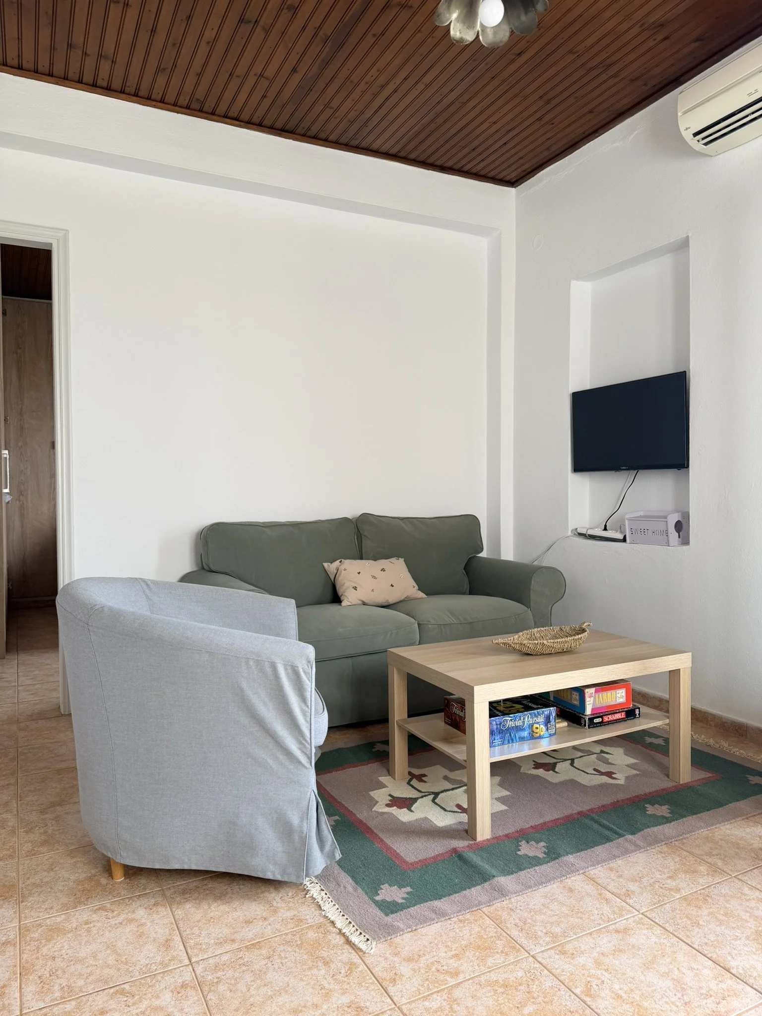 Living room with a gray armchair, green sofa, wooden coffee table with board games, a television mounted on white wall, and ceiling with wooden paneling.