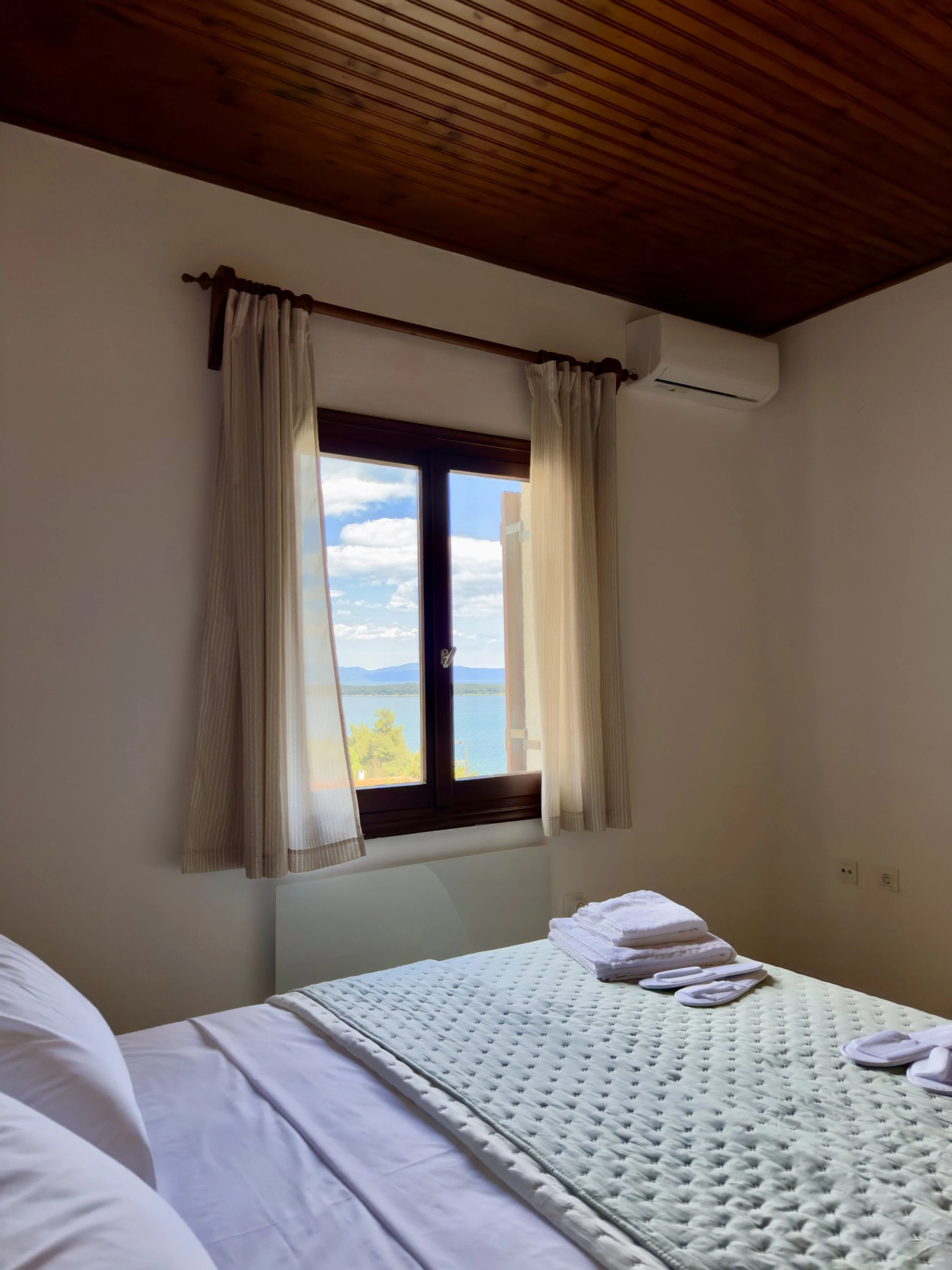 “Bright and cozy bedroom with a sea-view window, wooden ceiling, air conditioning, and neatly folded towels on the bed.”