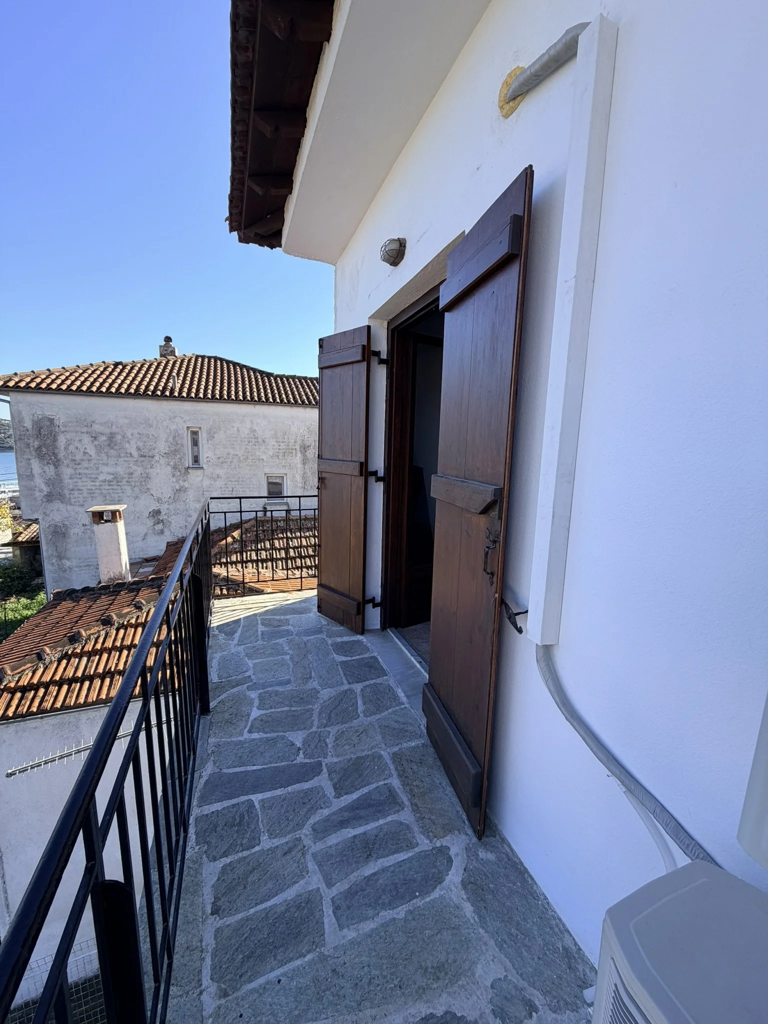 Small balcony with stone-tiled flooring, black metal railing, and white exterior wall with an open wooden window shutter. The view shows neighboring rooftops with orange tiles, an outdoor wall-mounted light fixture, and a building with a chimney set 