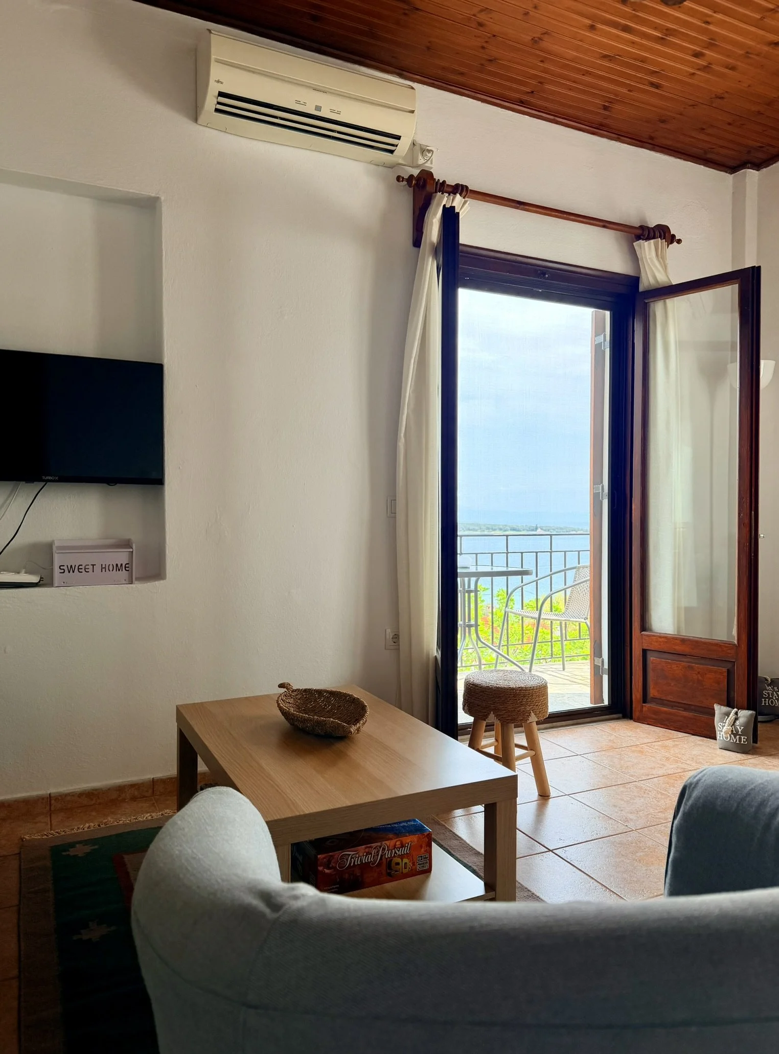 A cozy living room scene with a wooden coffee table, a grey armchair, and a view outside through an open door and window showing a balcony with white chairs and a view of the water and sky.