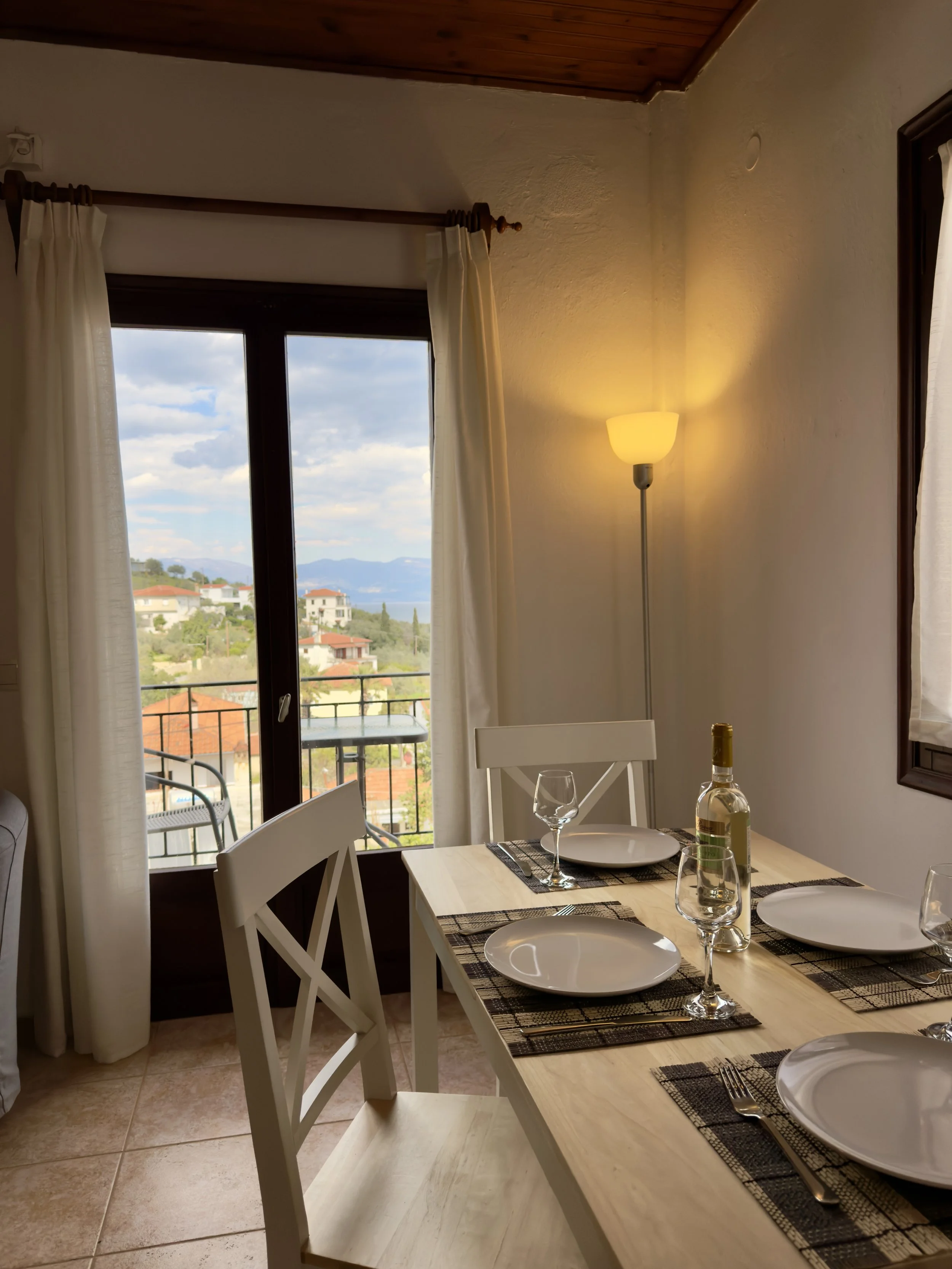A dining room with a table set for four, featuring white plates, wine glasses, and a bottle of wine. There are white chairs around the table, and windows with white curtain panels, revealing a view of houses, trees, and mountains outside. A warm yellow wall lamp provides soft lighting in the room.