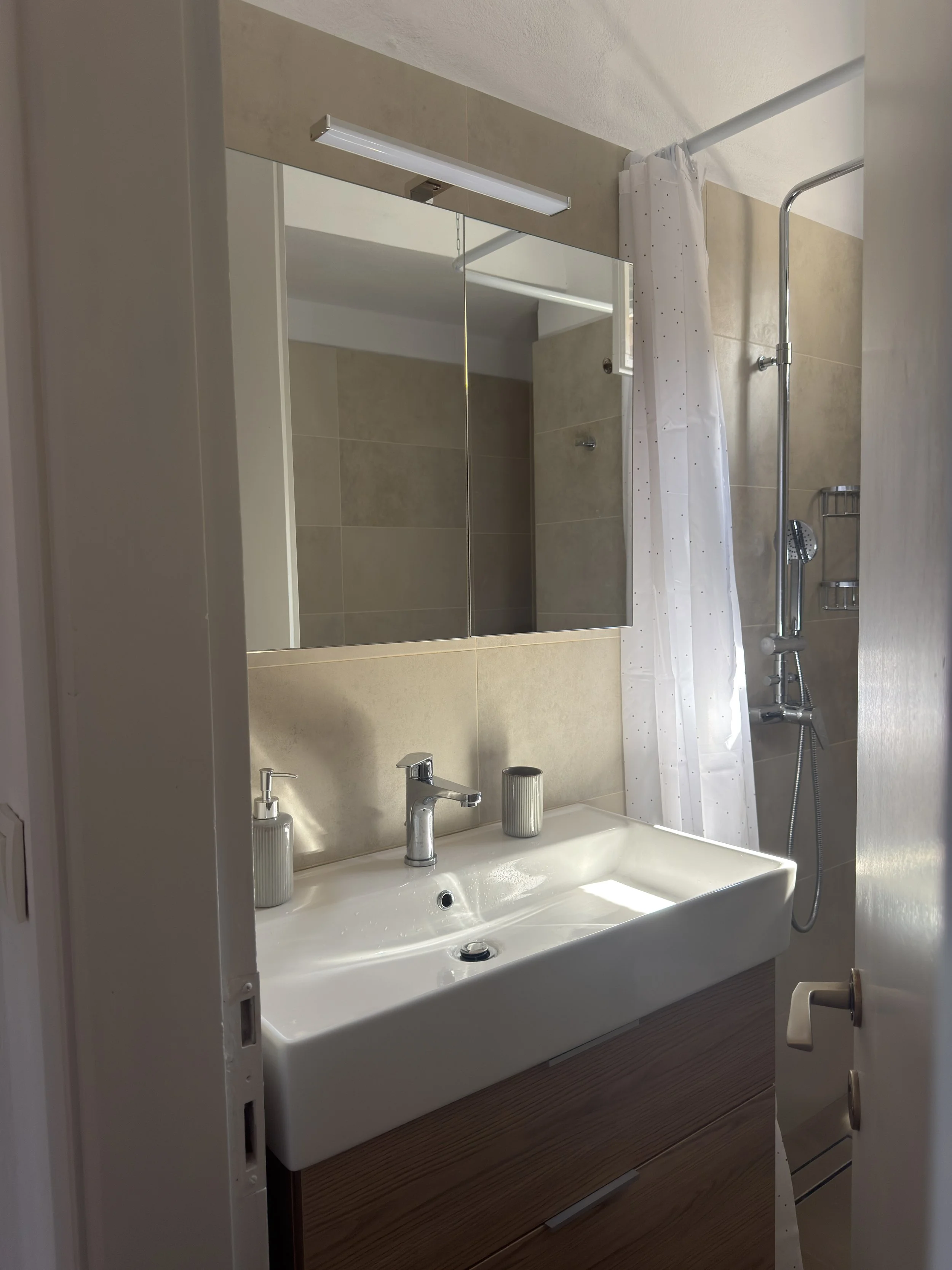 A bathroom with a white sink, two silver faucets, a mirror above the sink, a shower with a curtain to the right, and beige tiled walls.
