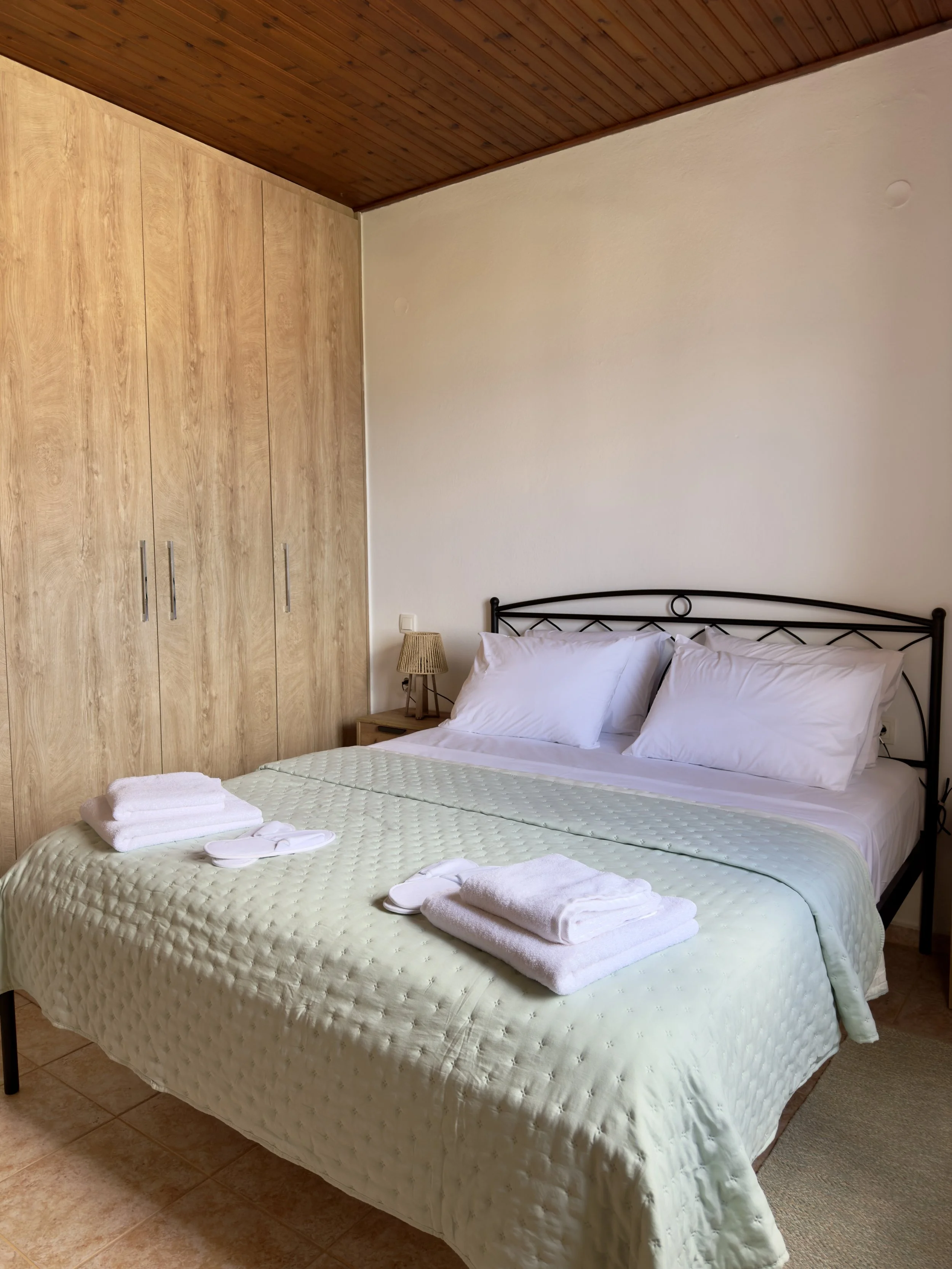 A neatly made bed with white pillows and a light green quilt, placed in a room with a wooden ceiling, beige wall, and a large wardrobe. There are two folded towels and slippers on the bed.