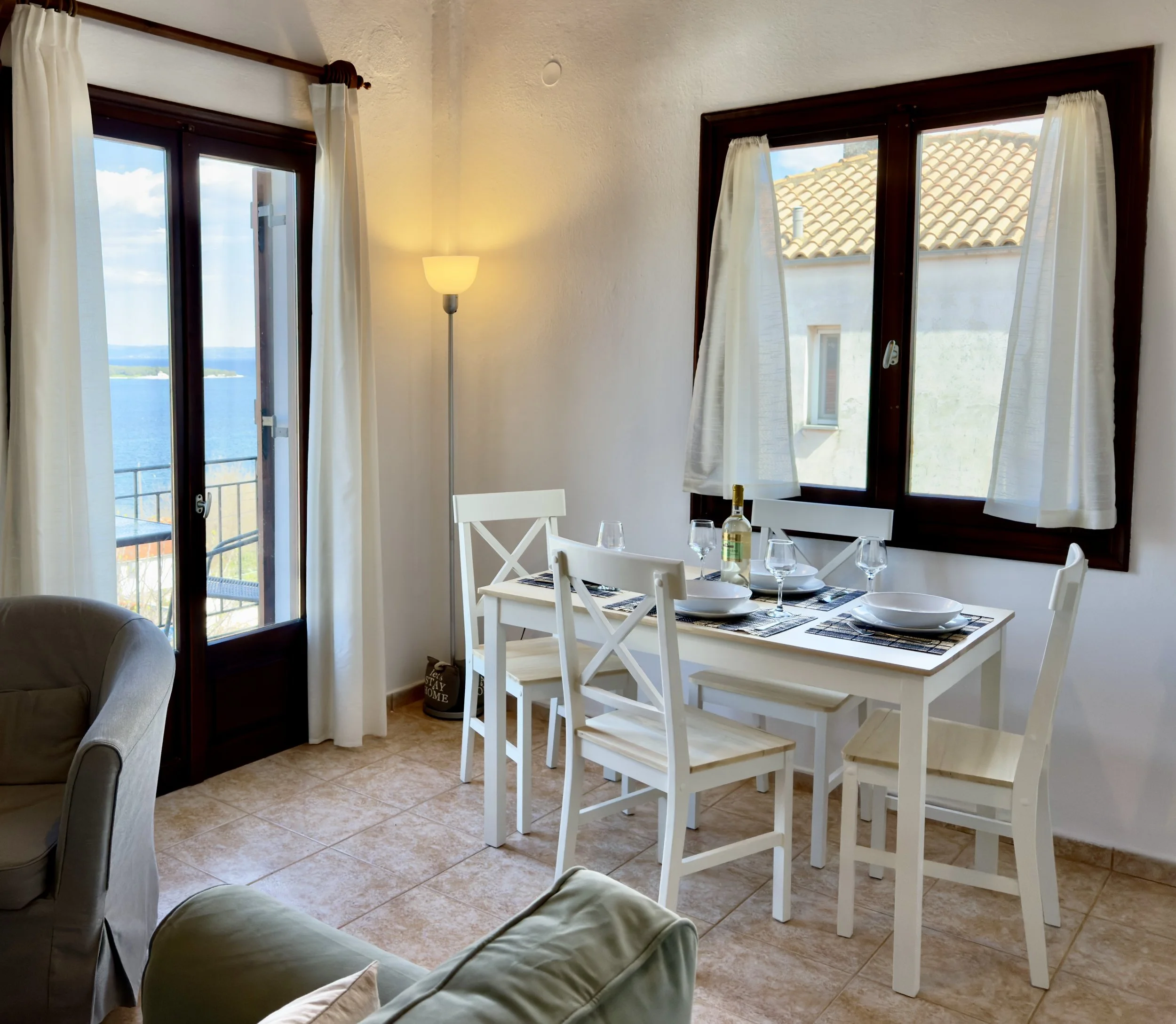 A dining room with a window view of water and sky. Inside, there is a white dining table with place settings and four white chairs. A corner lamp provides warm lighting, and beige tiled flooring completes the room's decor.