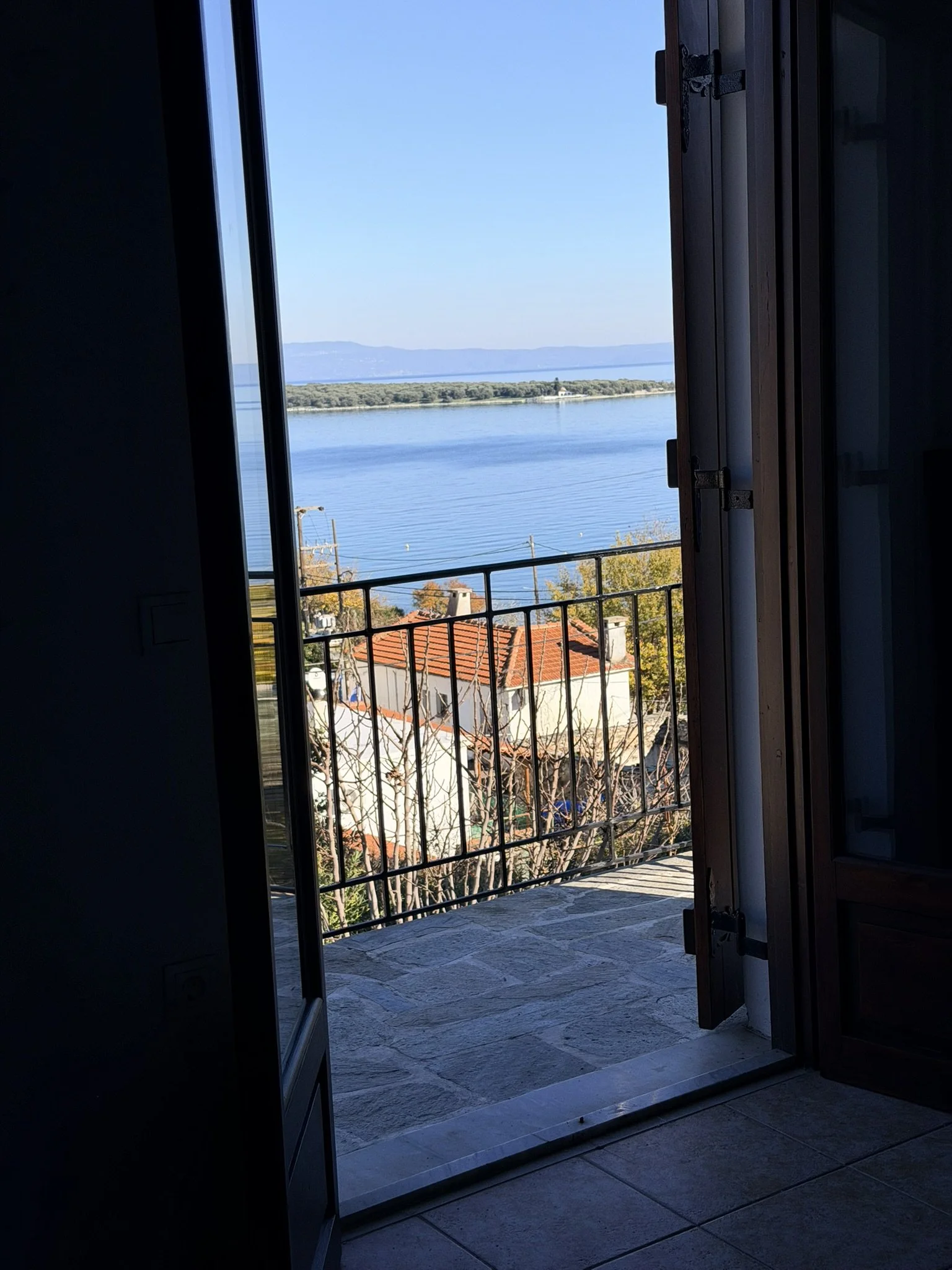 View from an open door or window showing a balcony overlooking a large body of water with land and distant mountains under a clear blue sky.