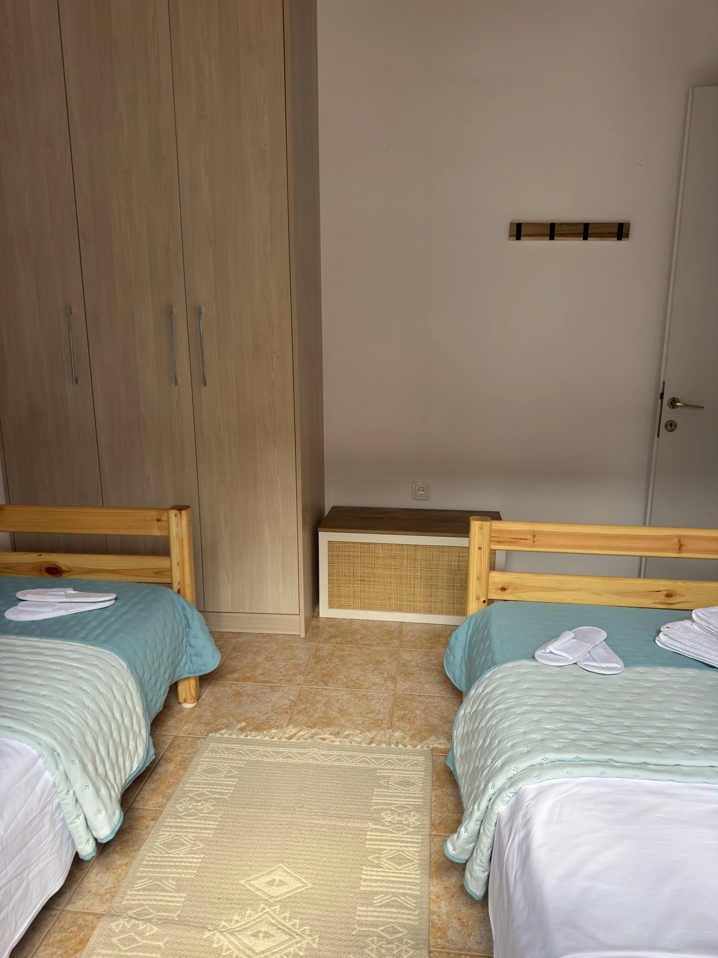 Two single beds with light blue quilted bedspreads and white pillows, towels, and slippers at the foot of each bed, in a room with wooden furniture, white walls, a beige rug, and a large wardrobe.